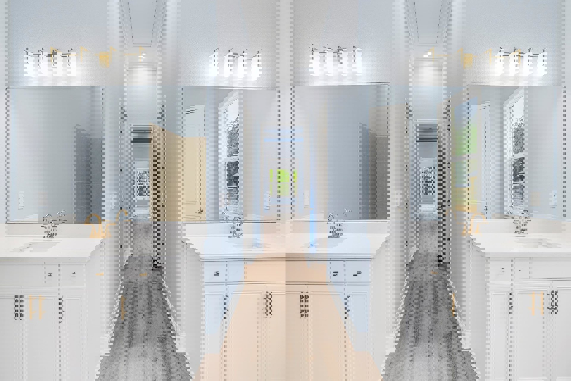 Bathroom with warm wood flooring, white vanity cabinet featuring gold handles, gold faucet, and white sink; partial view of white door and gold-framed rectangular mirror.