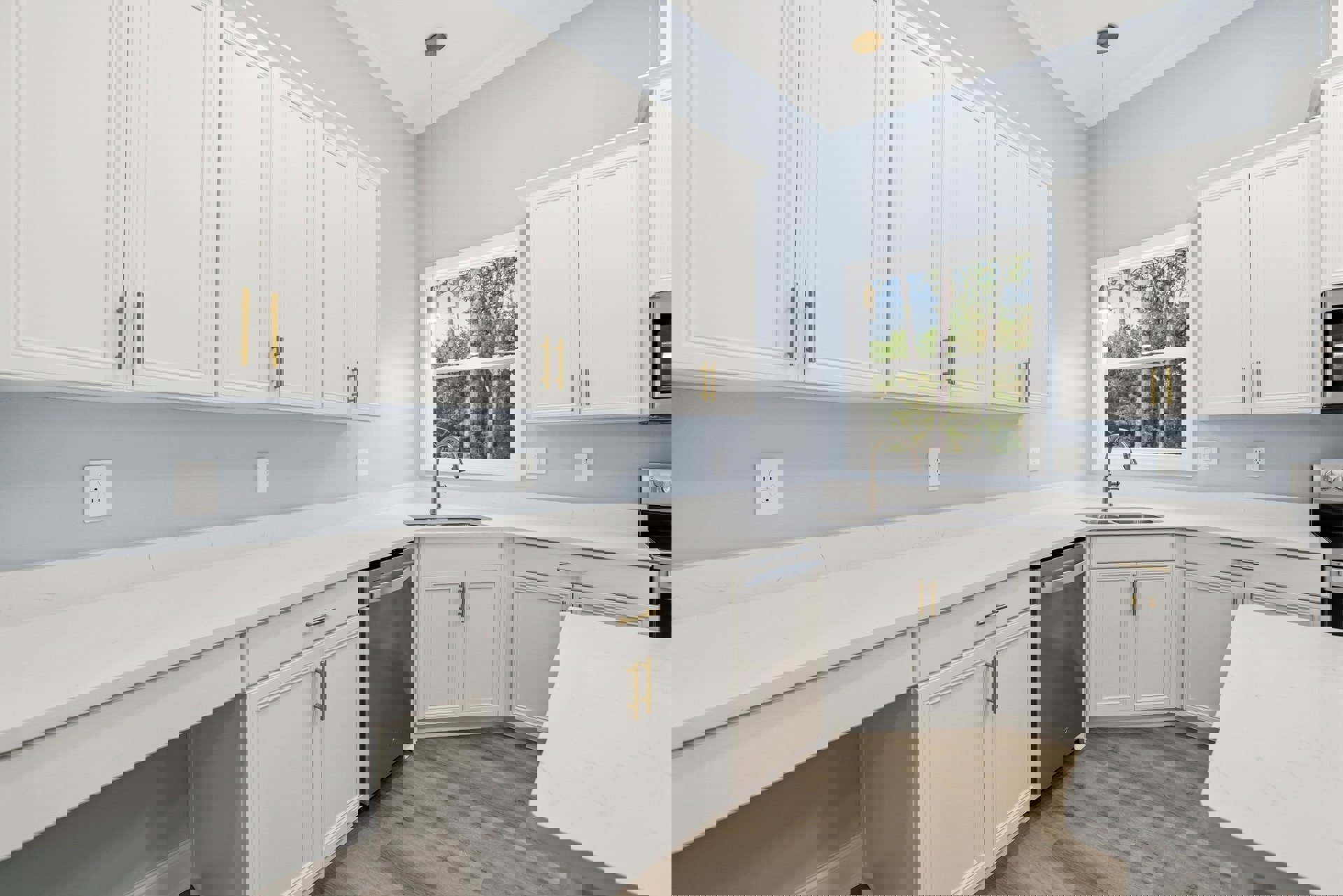 White kitchen cabinets with brushed metal handles, quartz countertop, stainless steel sink beneath a window showing green trees, white tile backsplash, and a white electrical