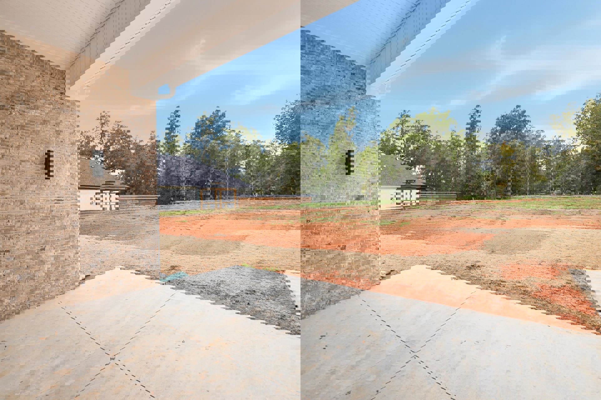 Red brick house with white roof, exterior wall-mounted light, concrete sidewalk bordered by dry grass, trees and cloudy sky in background
