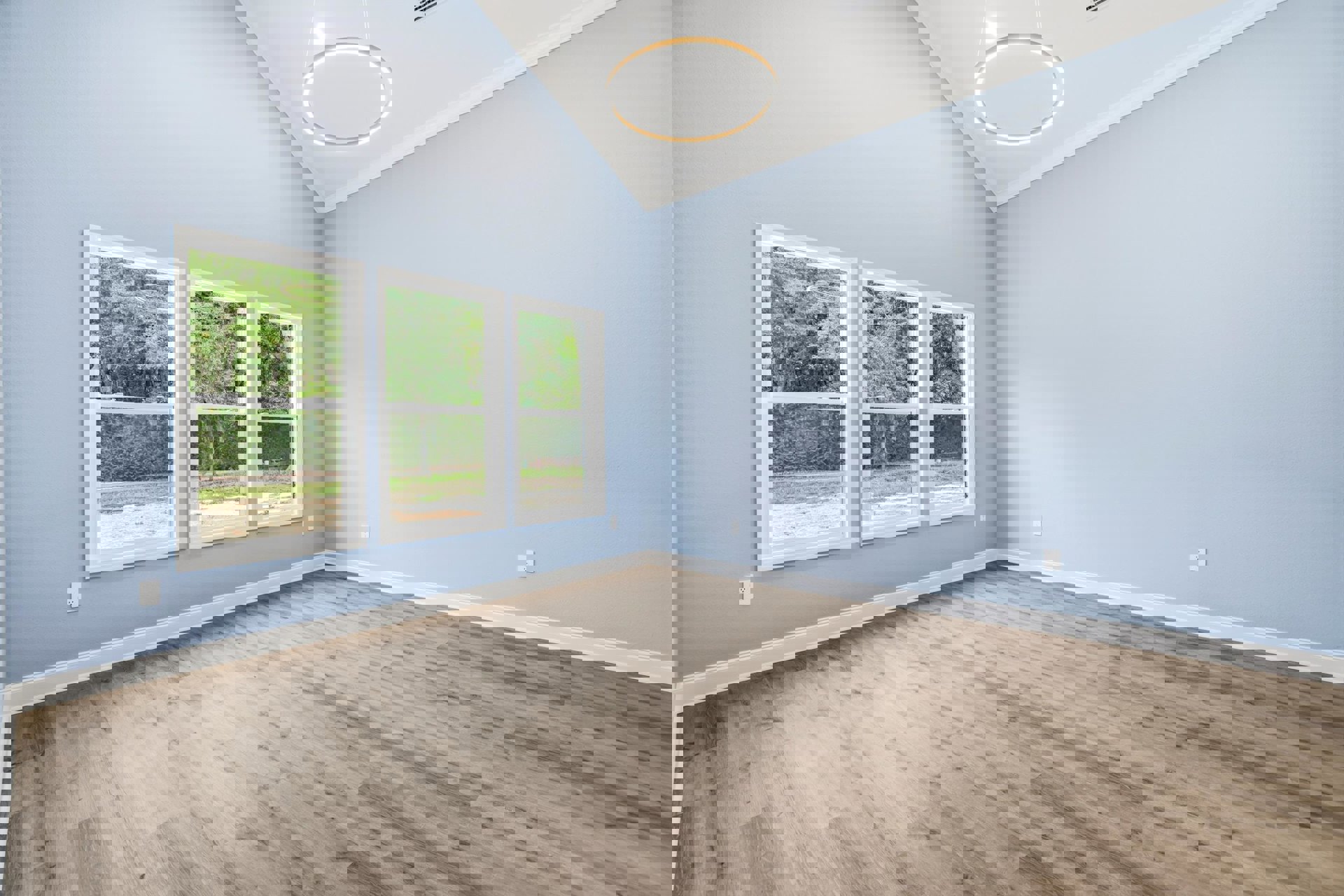 Spacious room with light wood flooring, large windows overlooking trees, white walls, and a ceiling light fixture