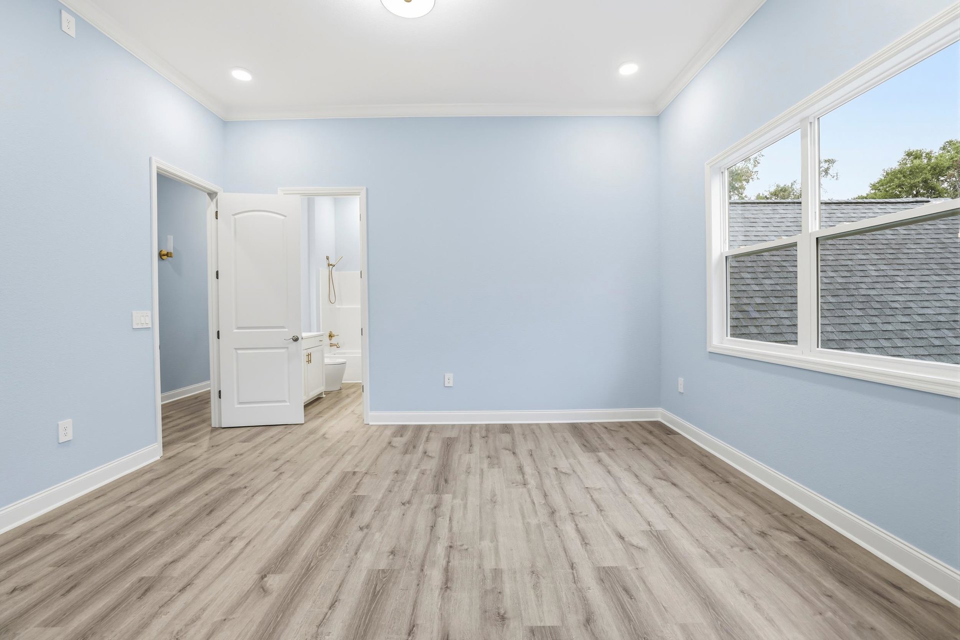 Wood flooring in a bright room with a white door, large window overlooking roof and trees, white plaster walls, and ceiling light fixture.
