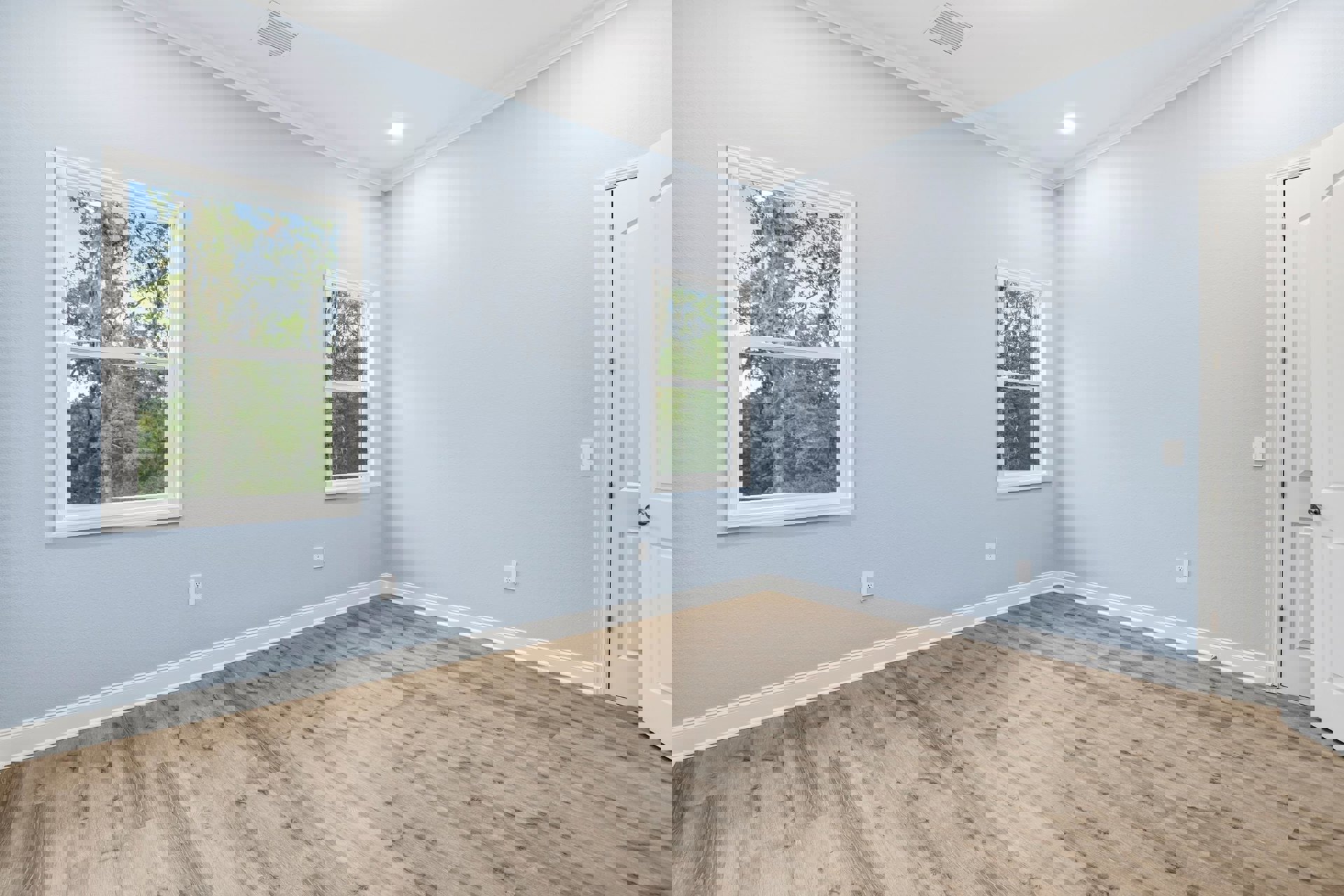Wood flooring with white baseboards, large windows showing trees outside, open white door, ceiling vents, and smooth plaster walls