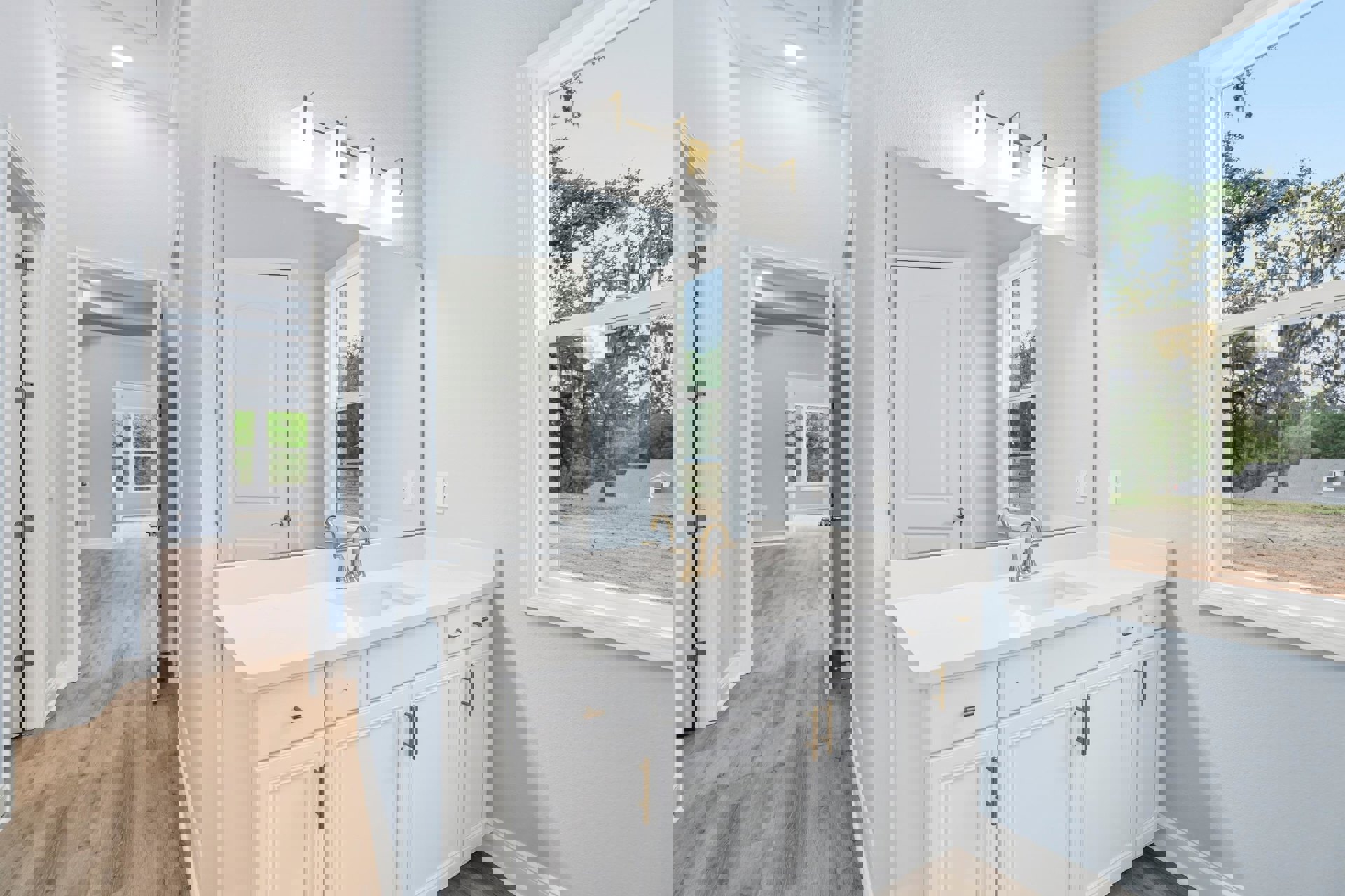 White bathroom with gold-handled sink, rectangular mirror above, four-bulb light fixture, white door with metal hinge, and tiled floor.