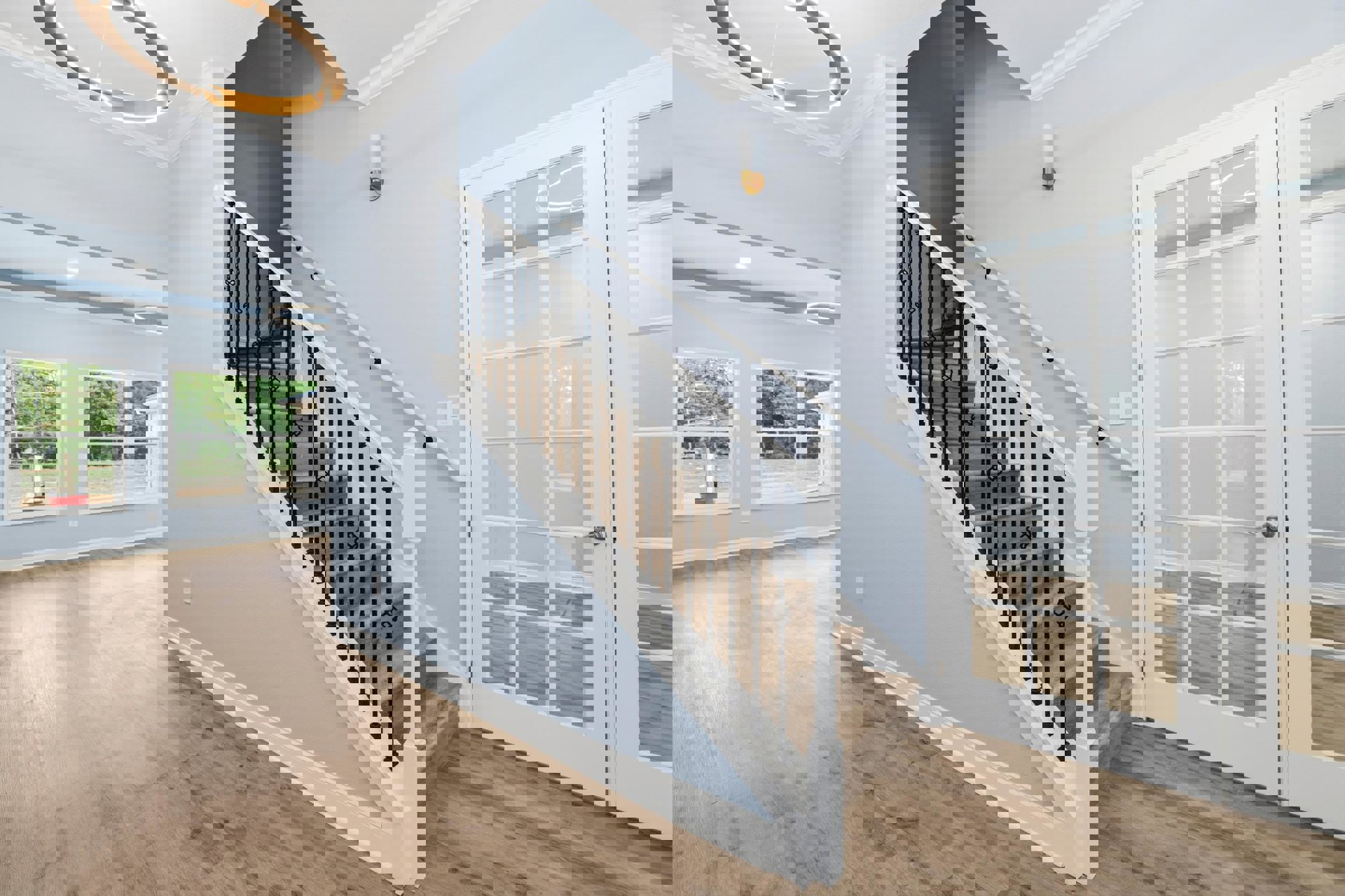 Open staircase with black metal railings, wood laminate steps, white walls, modern light fixture overhead, large window showing leafy trees, and white glass-paneled door.