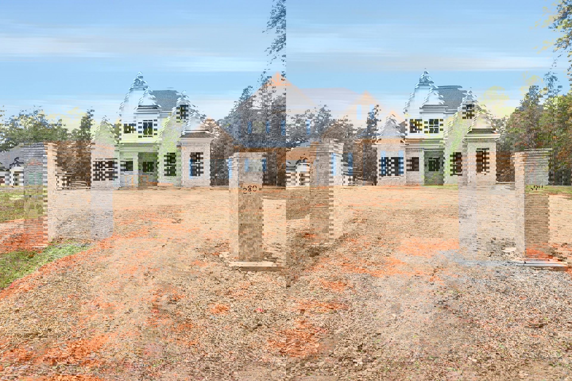 Two-story house with blue shutters, brick pillars marking the entrance, fenced yard, and mature trees under a clear blue sky
