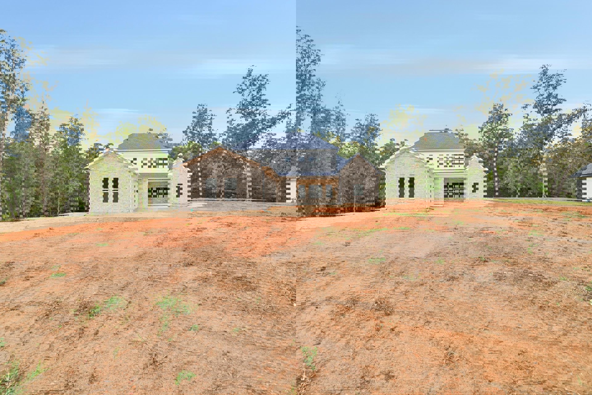 Two-story house with blue roof and white siding, surrounded by trees, set behind a large bare dirt lot under partly cloudy sky