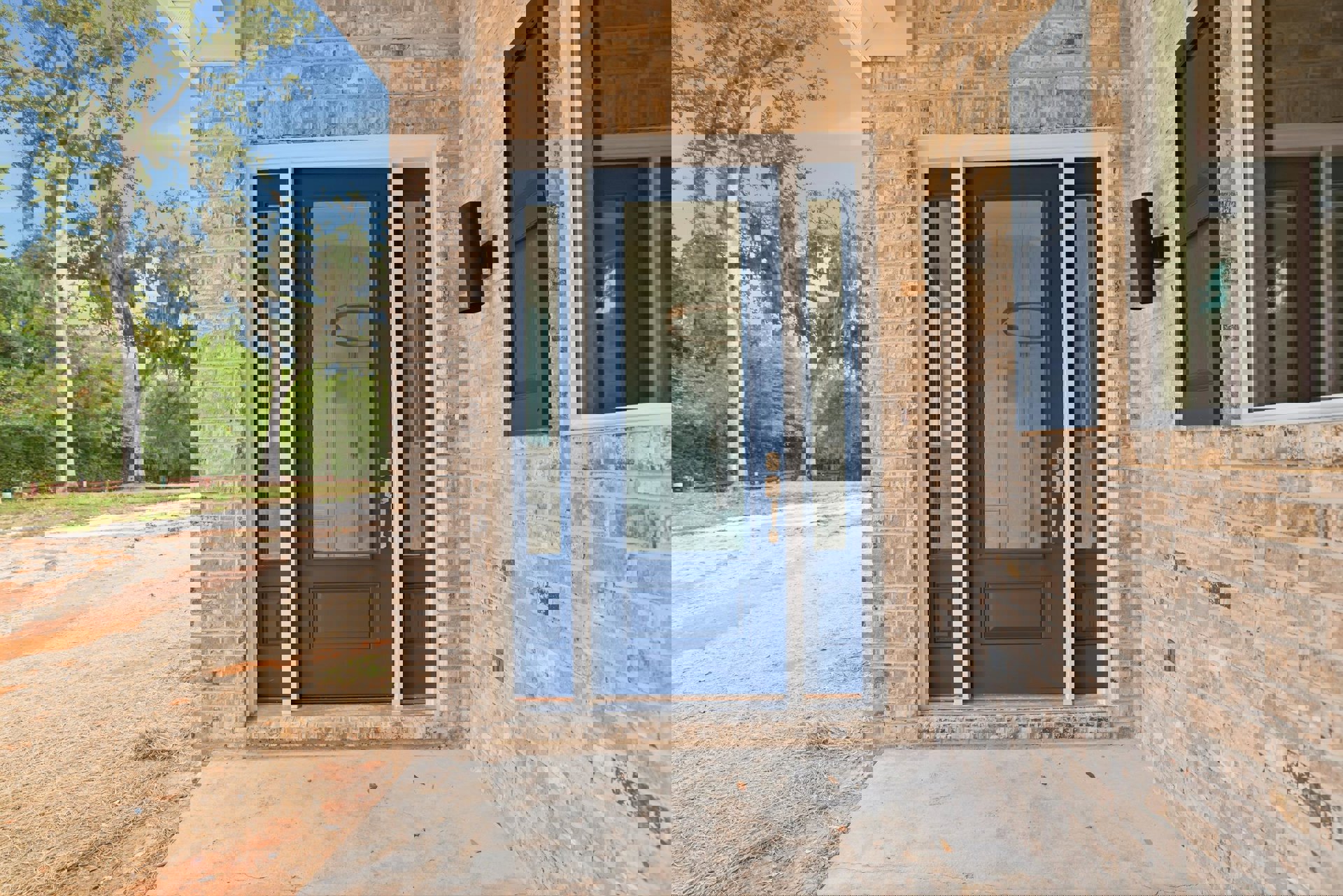 Blue front door with glass panel set in red brick exterior, surrounded by mature trees with hanging moss, concrete walkway, and grassy dirt road; window and wall details visible.