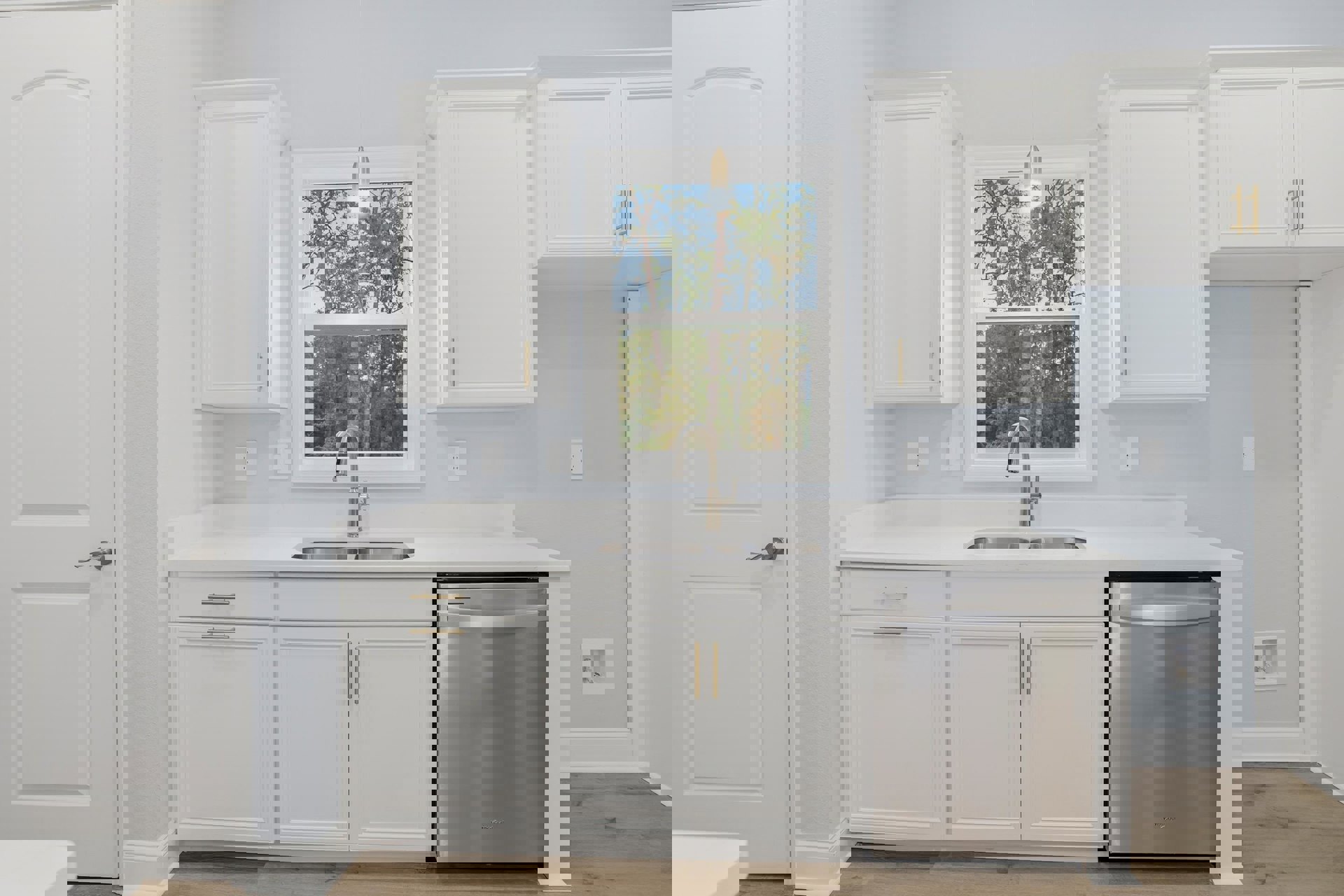 White kitchen cabinets, stainless steel sink under a window with trees outside, silver dishwasher, white doors with silver handles, light countertops.