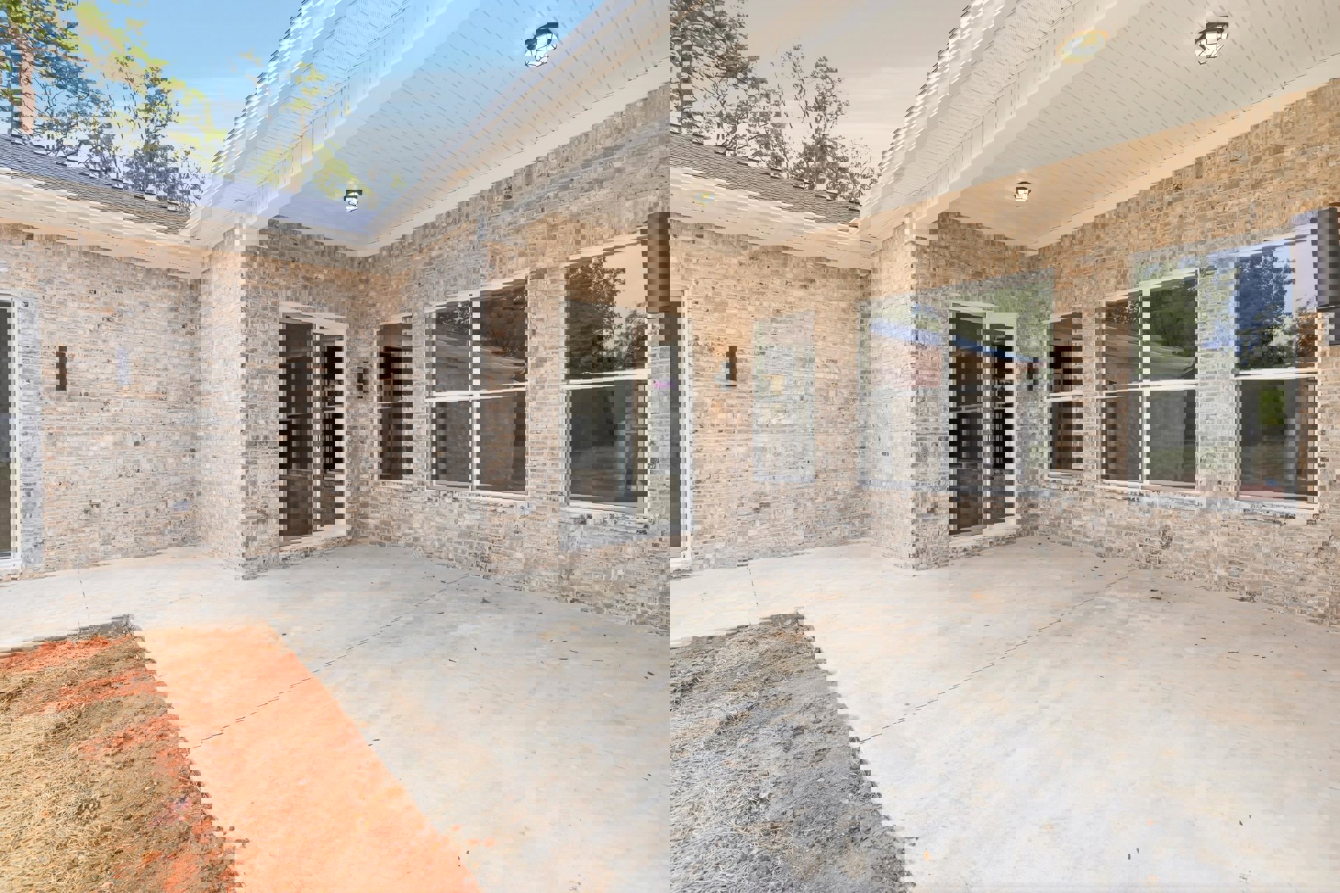 Red brick house with white roof, sliding glass door, white-framed windows, ceiling light fixture above covered patio, outdoor space bordered by trees.