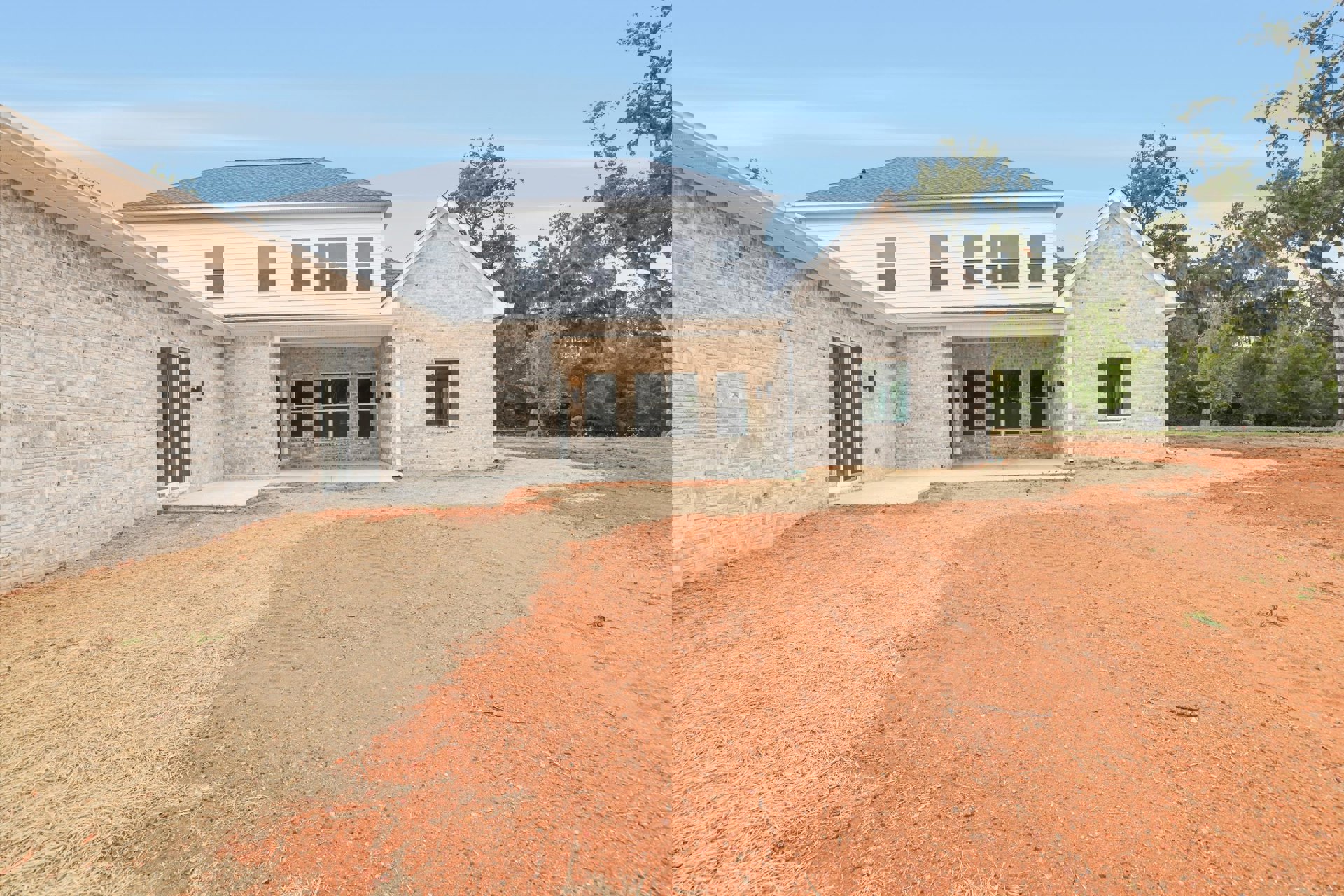 White house with brick accents, screened side window, dirt driveway, and surrounding trees under a clear sky