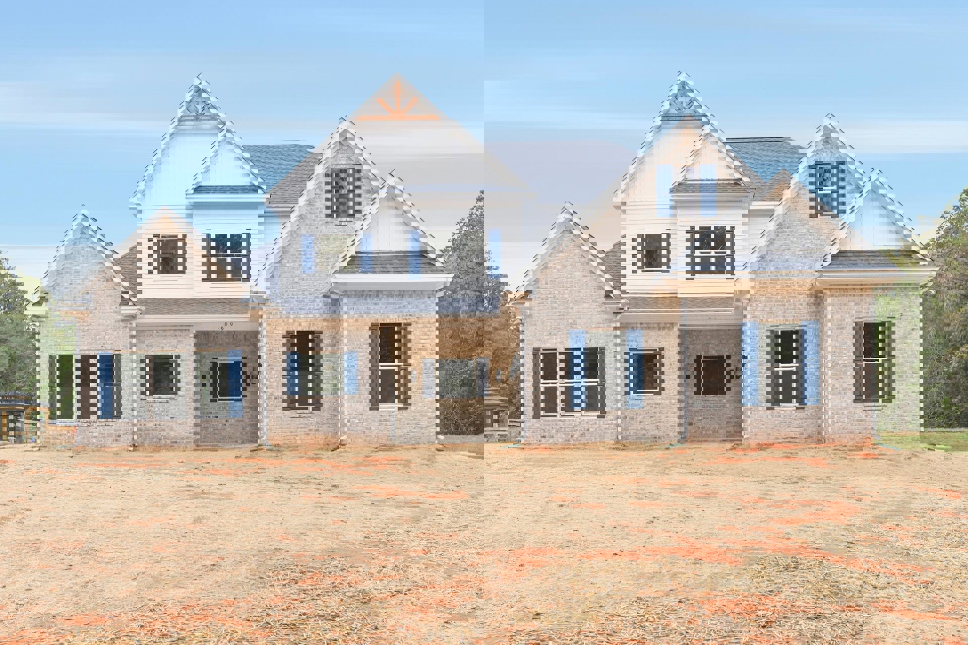 Two-story house under construction with blue shuttered windows, white trim, and white roof; dirt yard covered with straw, concrete driveway, and mature green tree in front