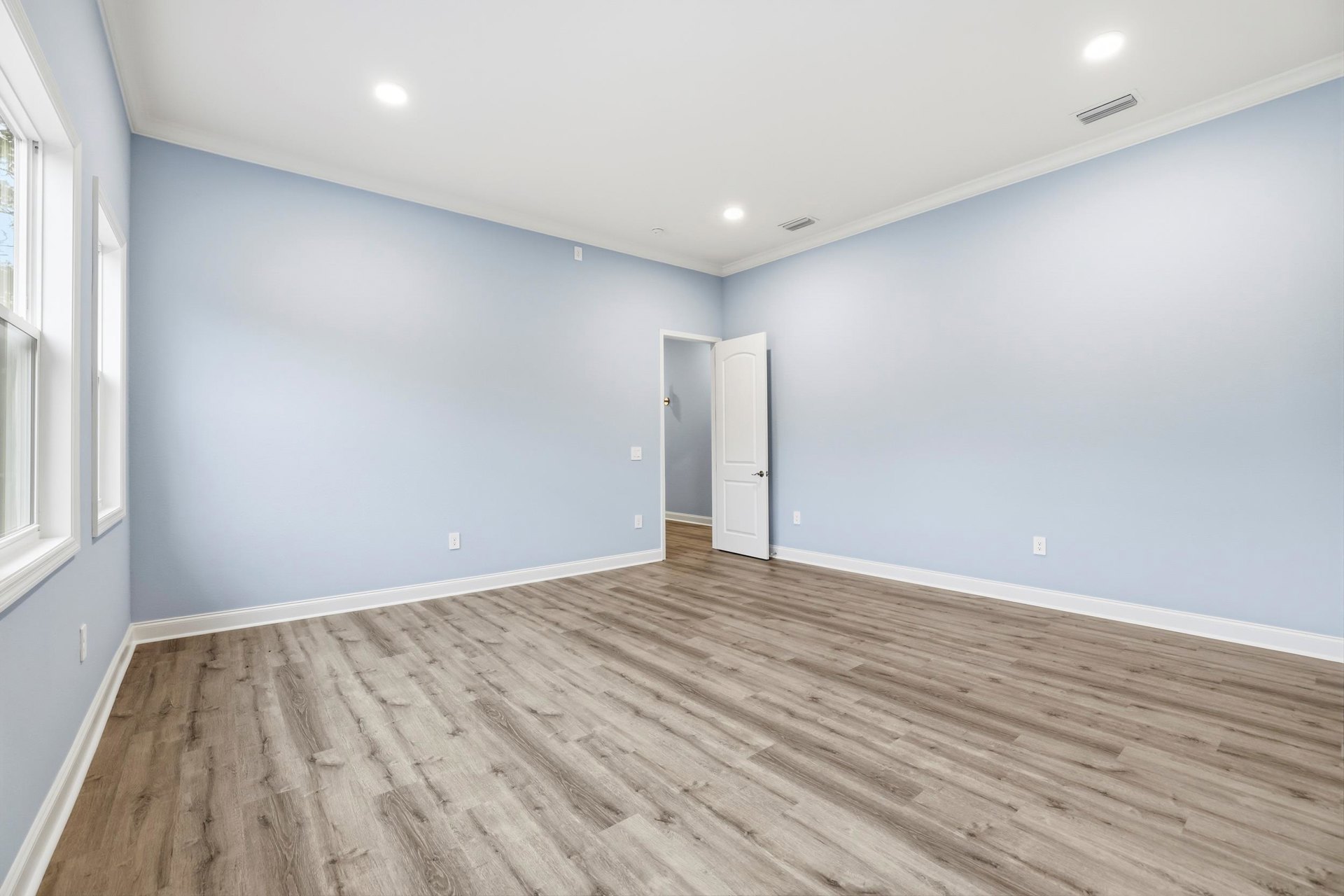 Wood flooring in a room with an open white door featuring a silver handle, white plaster walls, ceiling vent, and a ceiling light fixture.