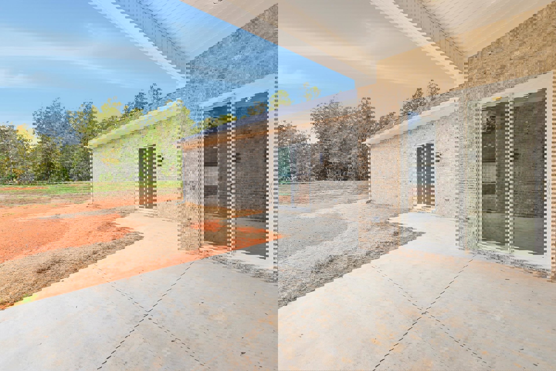 Red brick exterior with sliding glass door and large window opening onto concrete patio bordered by dirt and grass, surrounded by trees under open sky