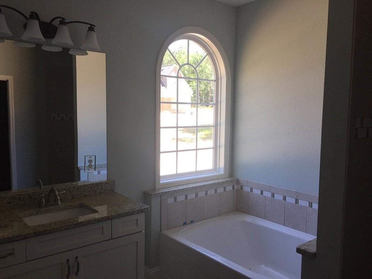 Freestanding white bathtub beside a large window with white trim, modern sink with chrome tap, light gray tile flooring, white cabinetry, and minimalist light fixture