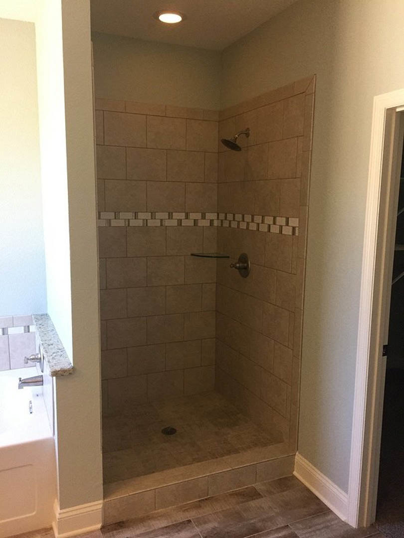 Walk-in shower with tile trim, chrome shower head and metal rod, glass door handle, built-in shelf, white light fixture, and plaster walls in a modern bathroom.