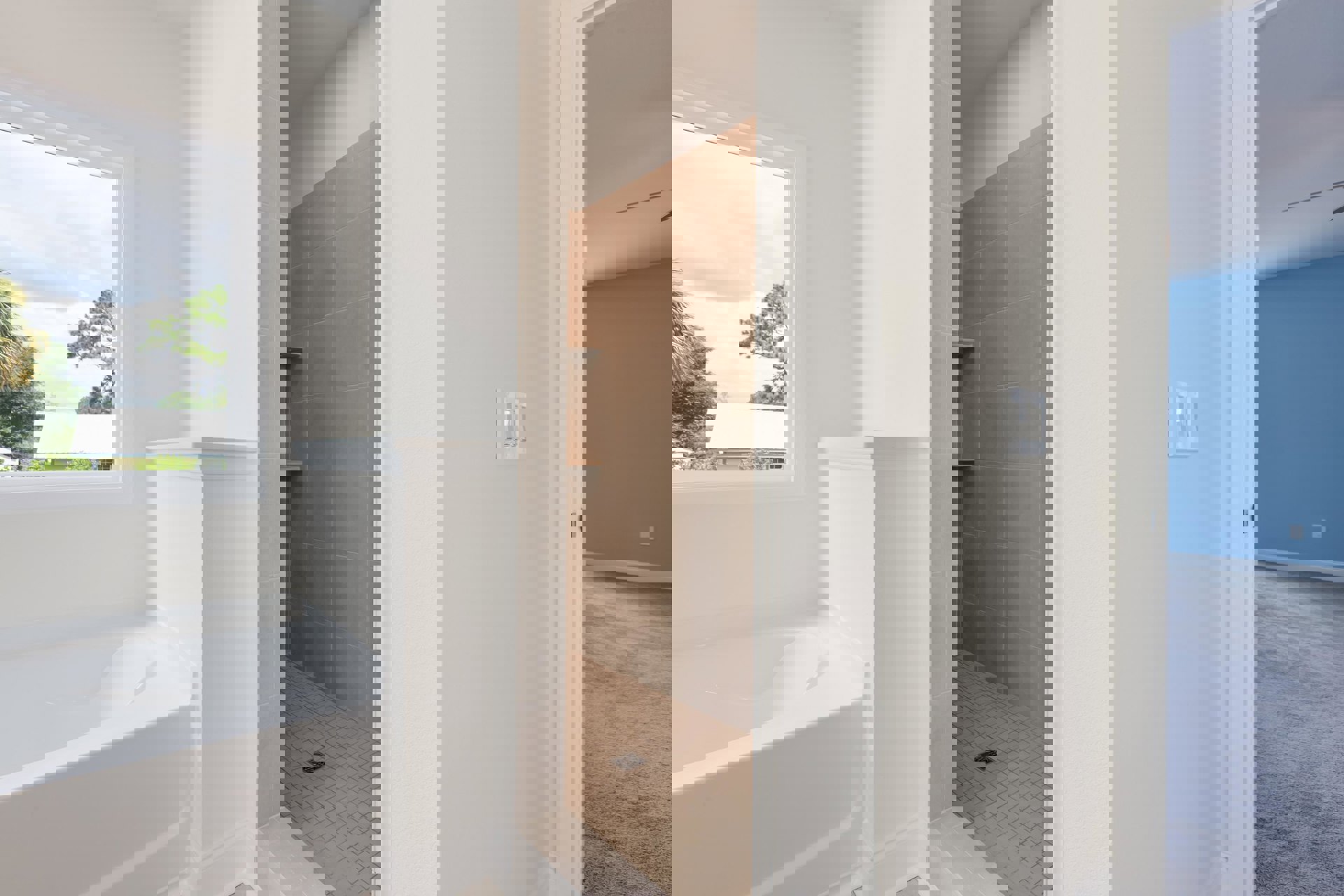 Bathroom with white curved-edge bathtub, glass-enclosed shower, light gray tile flooring, window overlooking trees, chrome plumbing fixtures, and row of white wall switches