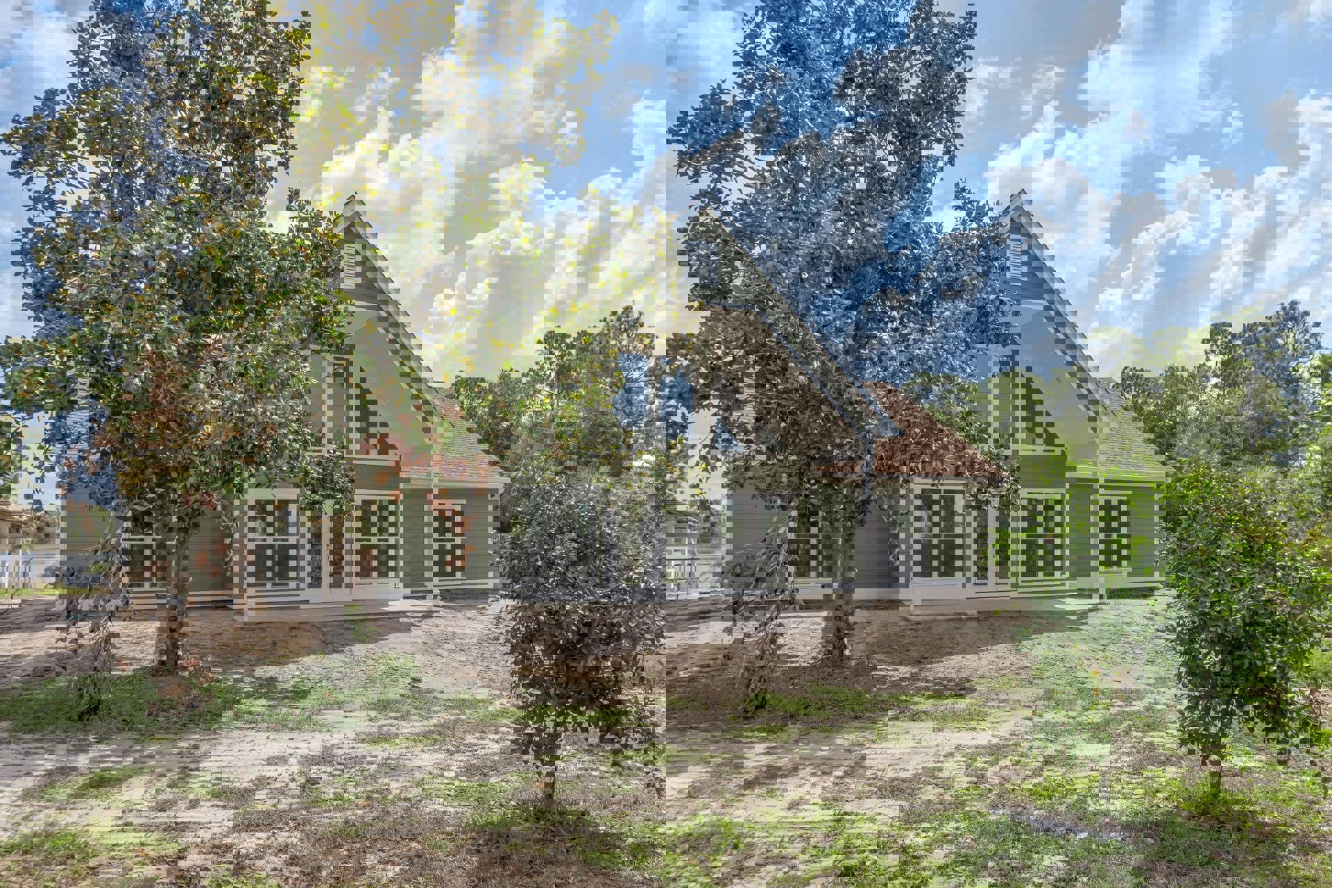 Two-story house with white-framed windows, light siding, dirt patch in front yard, mature trees behind and beside the home, cloudy sky overhead