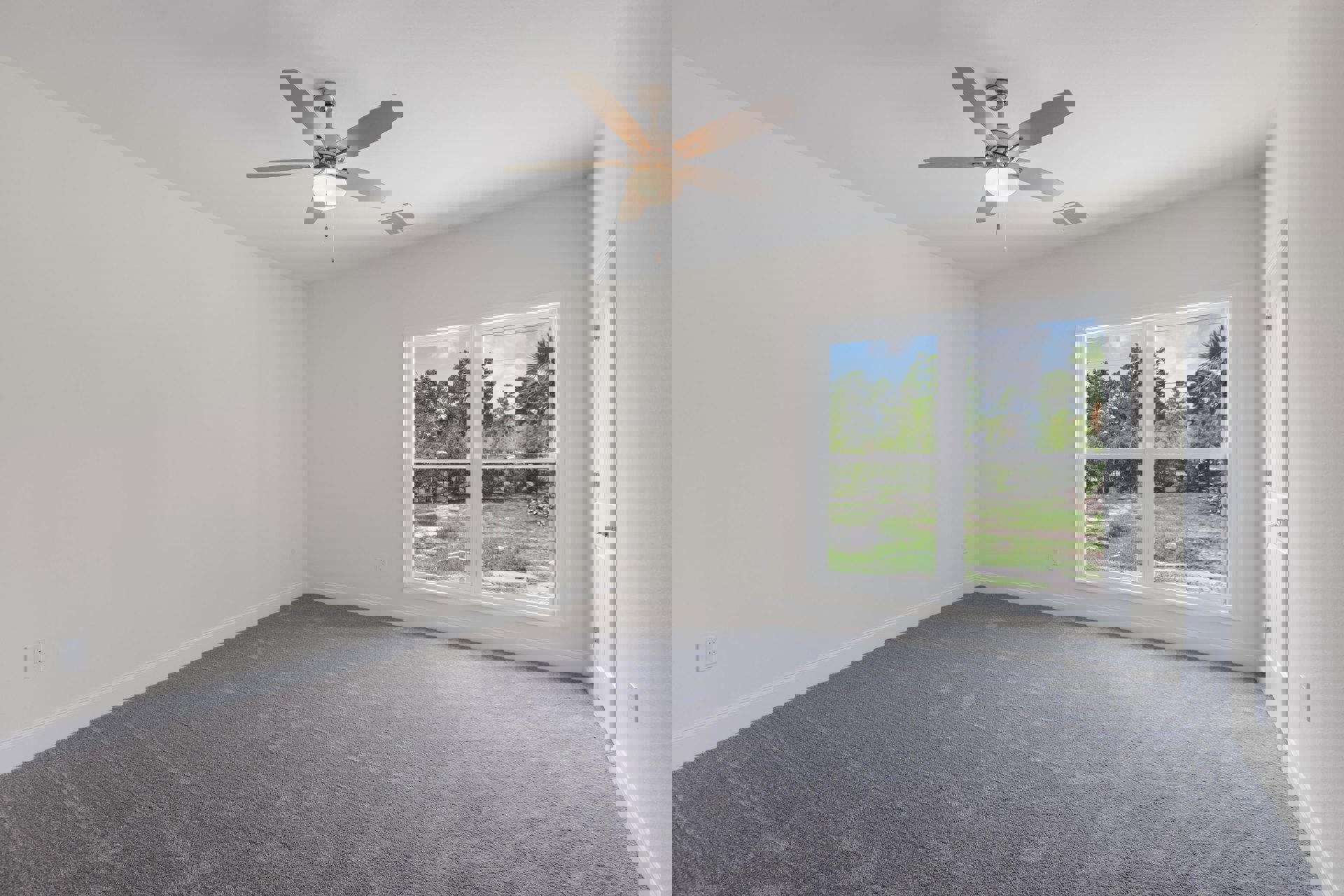 Carpeted room with white walls, ceiling fan with light fixture, large window showing trees, grass, and fence outside