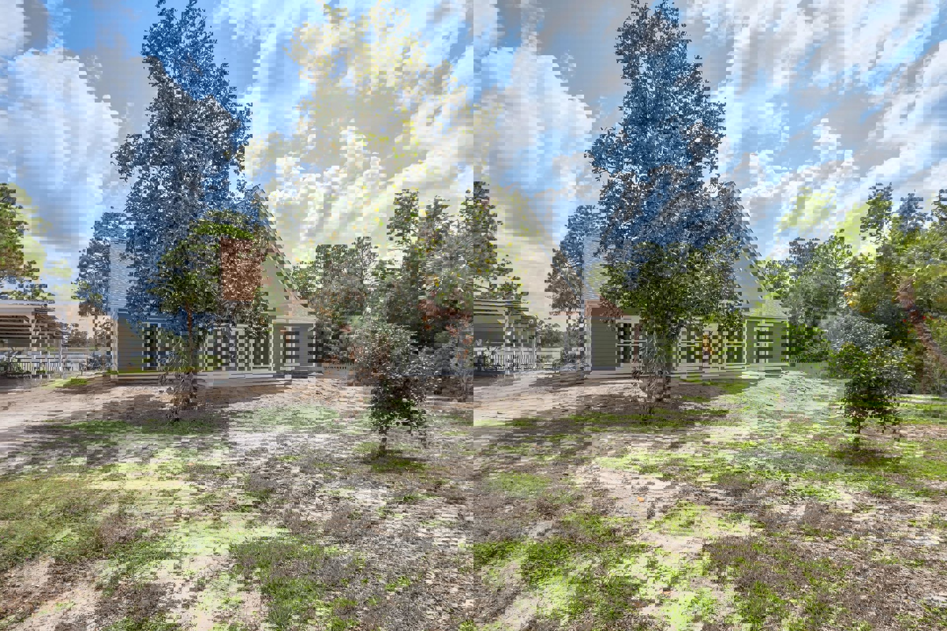 Modern house with light siding and metal railing, set behind a spacious grassy yard with a large mature tree and cloudy sky overhead