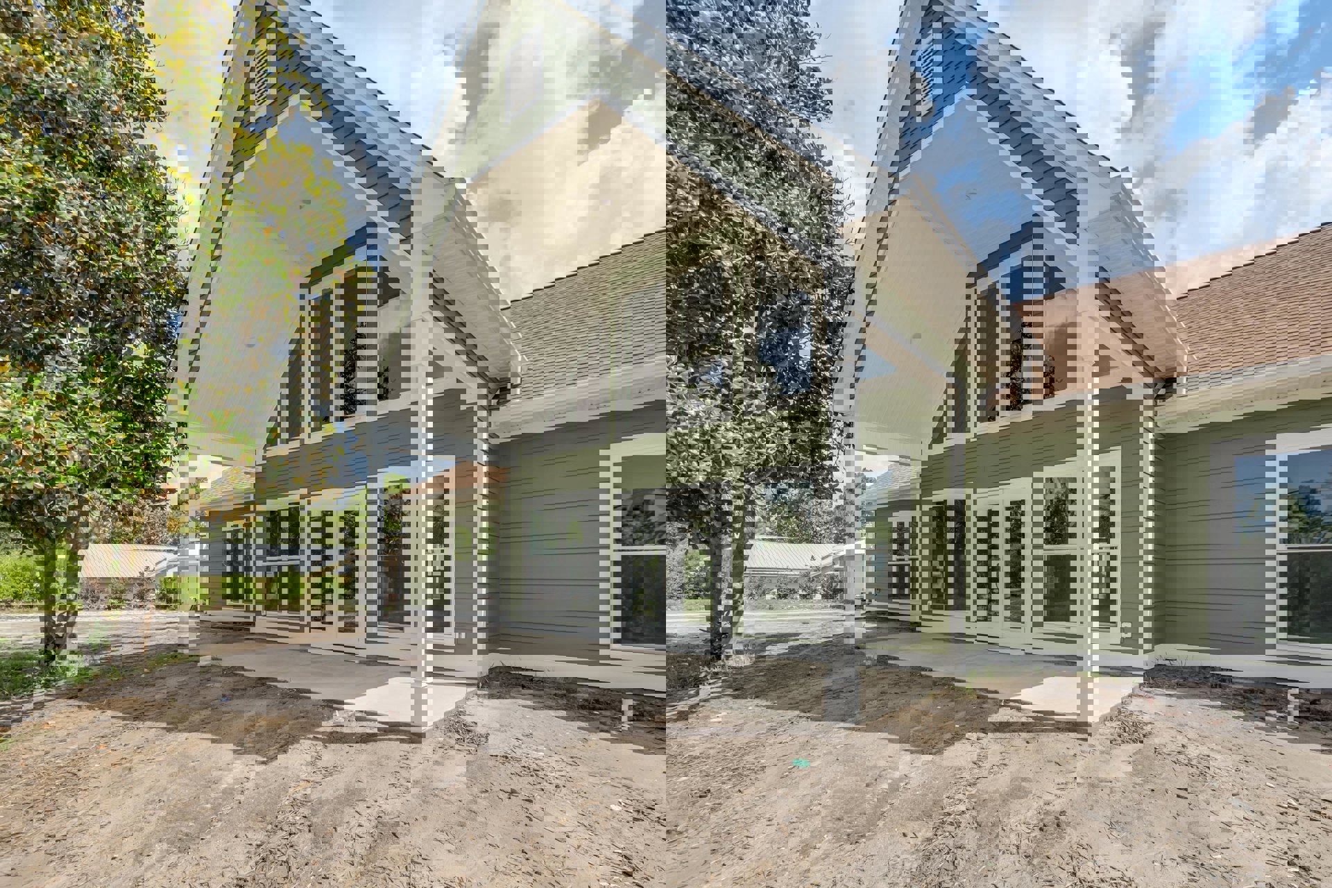 Spacious front porch with white columns, green metal roof, large windows reflecting leafy trees, white exterior vent, mature tree with dense foliage in foreground