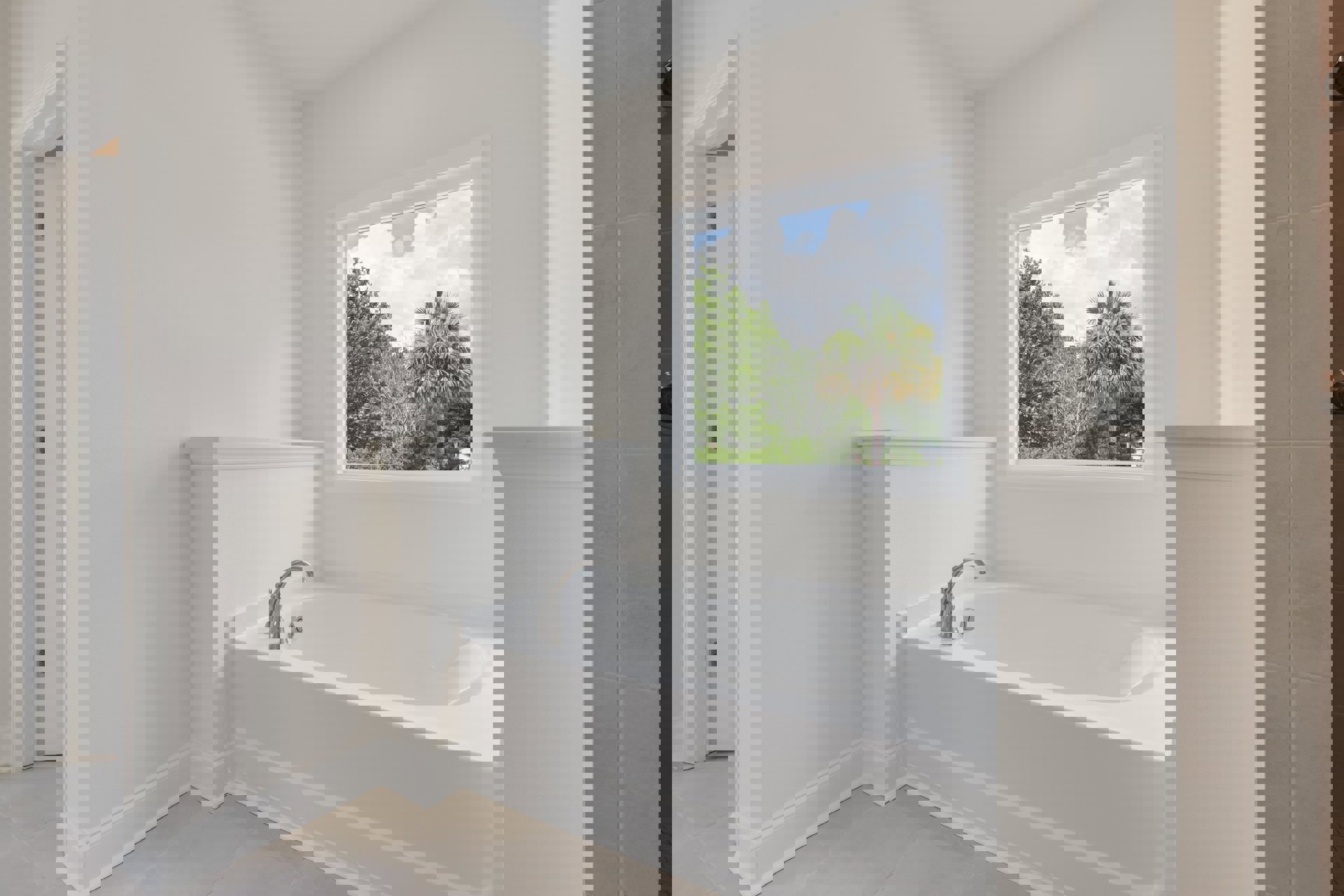 Freestanding white bathtub with silver faucet beneath large window, bathroom walls tiled in light gray, view of trees and blue sky outside