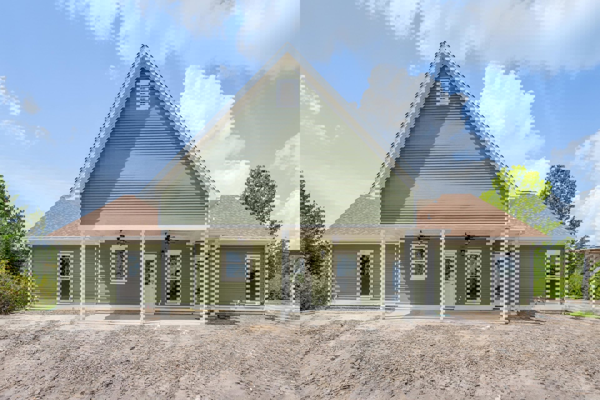 Green house with white trim and pillars, white-framed windows, white vent, porch, dirt ground with scattered sticks, illuminated window, cloudy sky, trees in background.