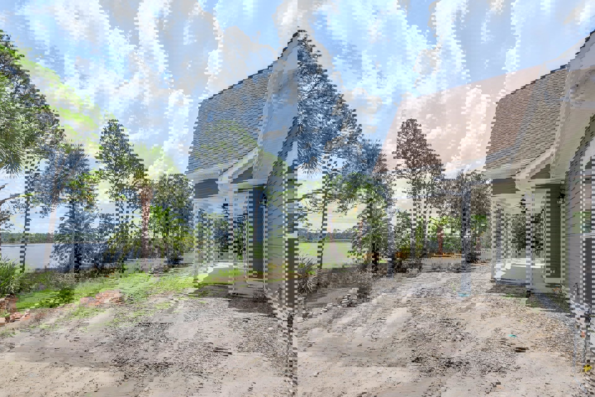 Two-story house with covered front porch, surrounded by mature trees, dirt road with scattered sticks leading to sandy yard area under partly cloudy sky