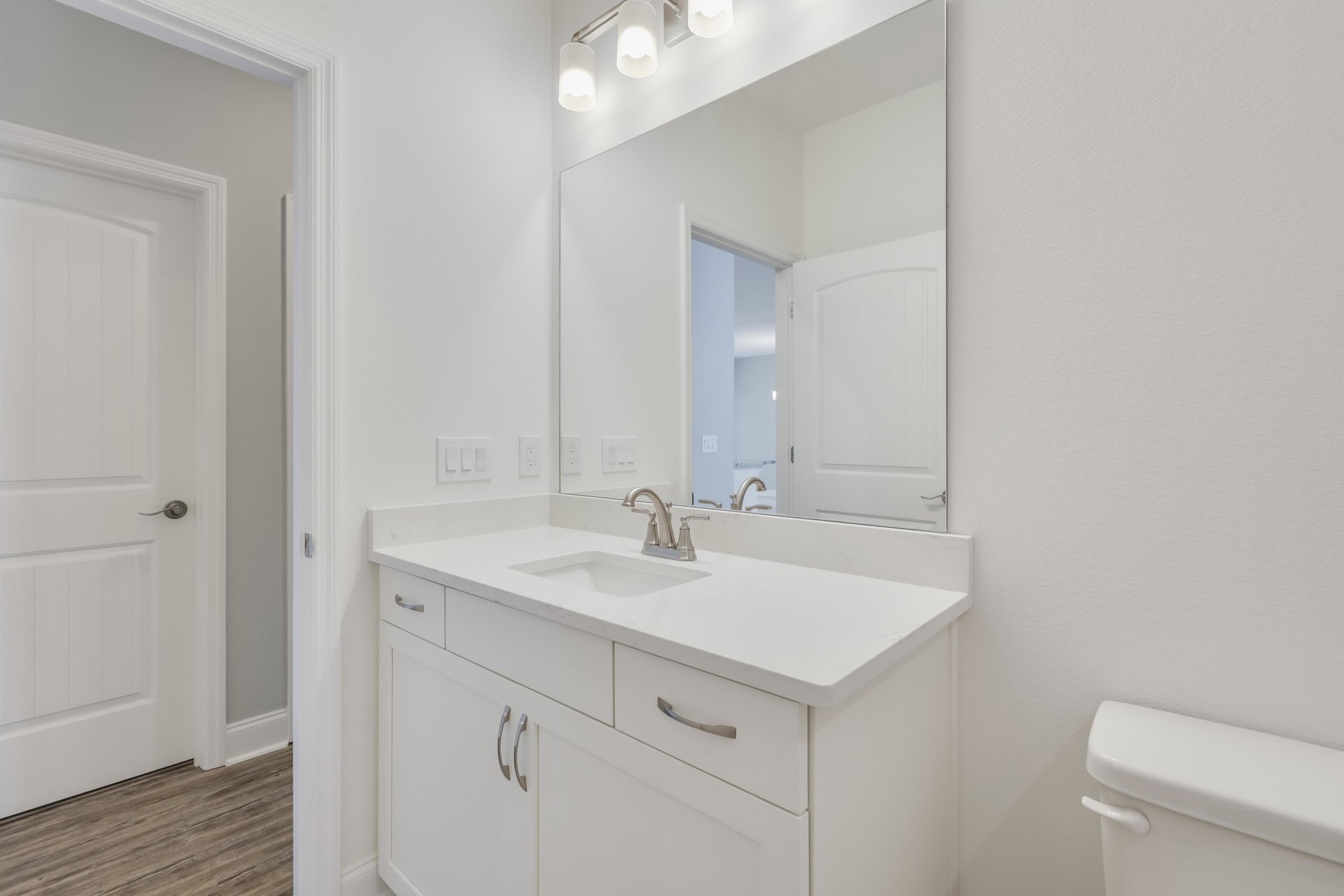 White bathroom with wood flooring, wall-mounted mirror above rectangular sink with silver faucet, white door with silver handle, and wall sconce lighting.