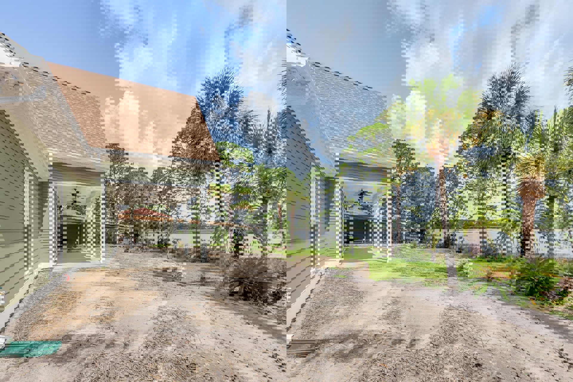 Covered porch with white railings overlooking a lake, dirt road bordered by grass and sticks, green bush near the house, gray shingle roof, green plastic container on ground