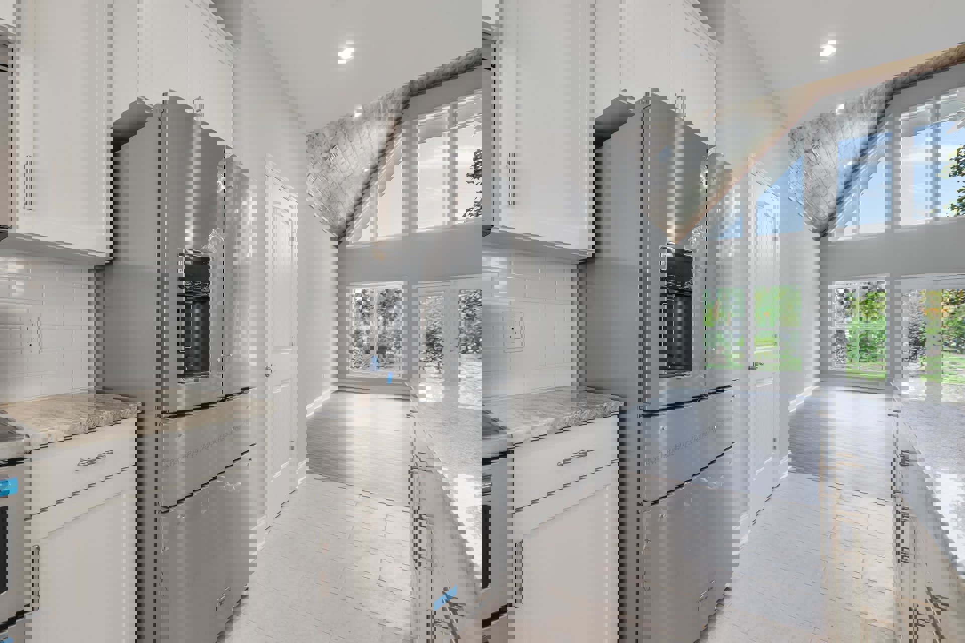 Stainless steel refrigerator with water dispenser beside white cabinetry, grey countertop, and window showing blue sky