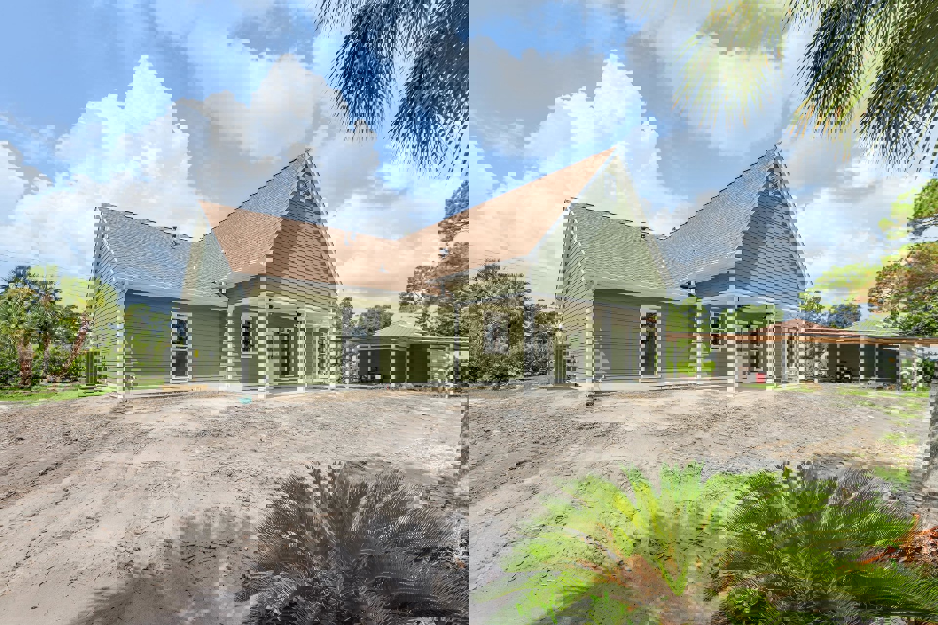 Single-story house with white roof and green exterior, surrounded by palm trees and dirt landscaping under a partly cloudy sky