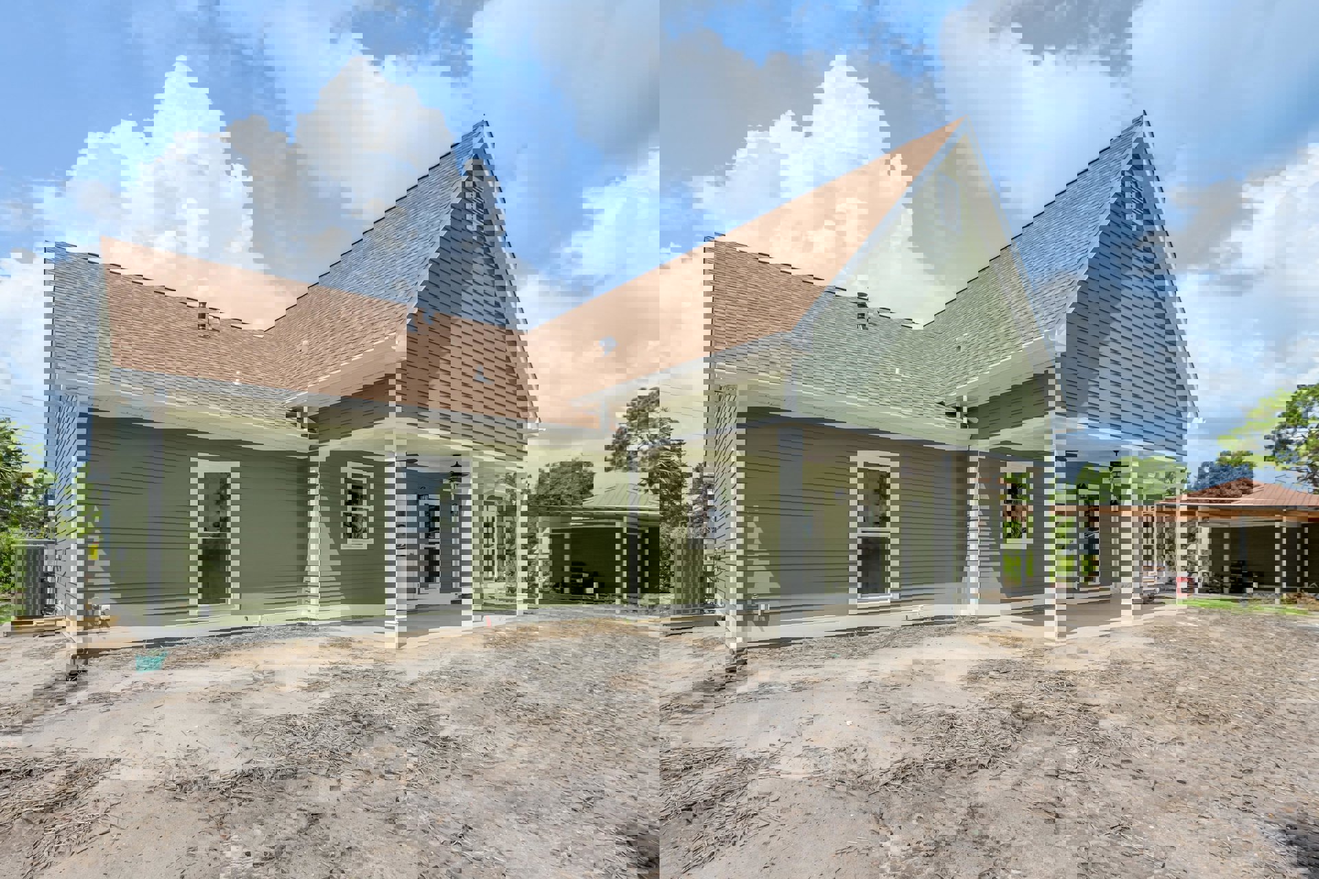 Green siding house with white framed windows, one featuring a dinosaur face decoration, white door, dirt and sticks covering the ground, blurred red container near porch, cloudy