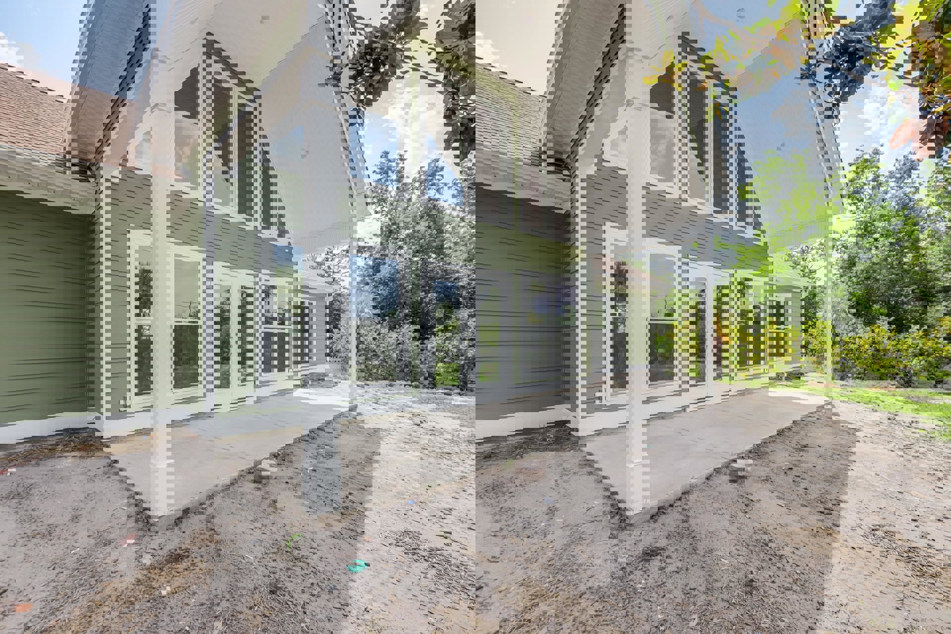 Modern home with white siding, glass-paneled double doors, large windows reflecting palm trees, covered patio area, dirt driveway, and sidewalk adjacent to the building