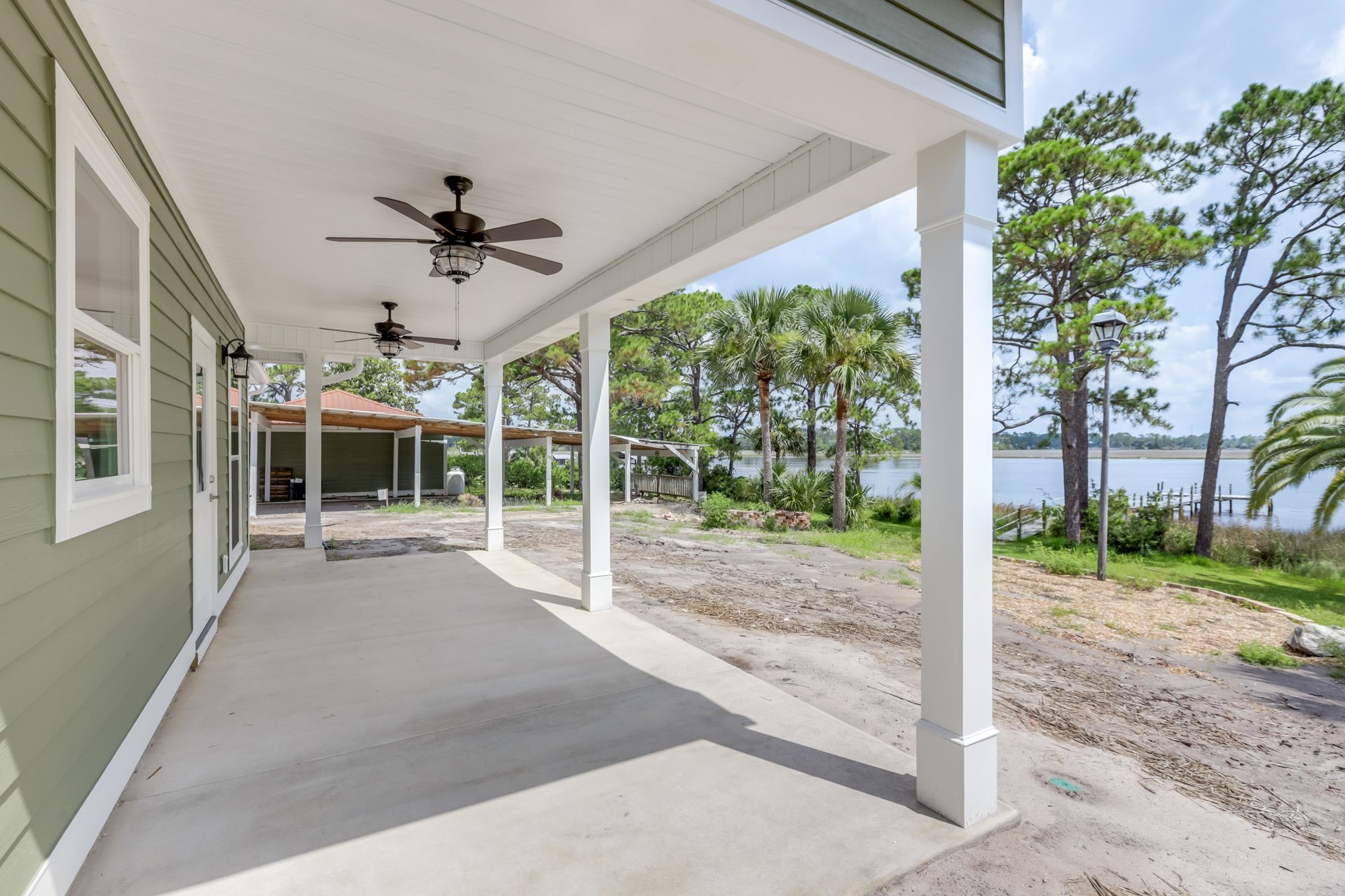 Covered porch with ceiling fan and light, concrete floor, white pillar displaying a sign, street lamp and leafy trees in background, sunlight casting shadows