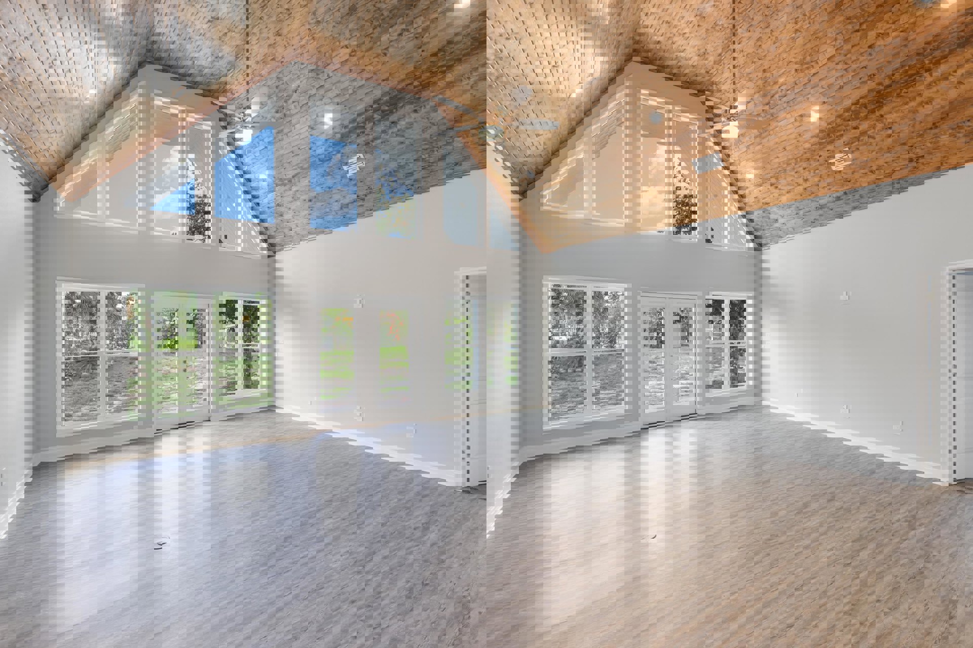 Spacious room with hardwood floors, ceiling fan, white door with metal hinge, large windows showing blue sky, clouds, and palm tree, wall vent visible