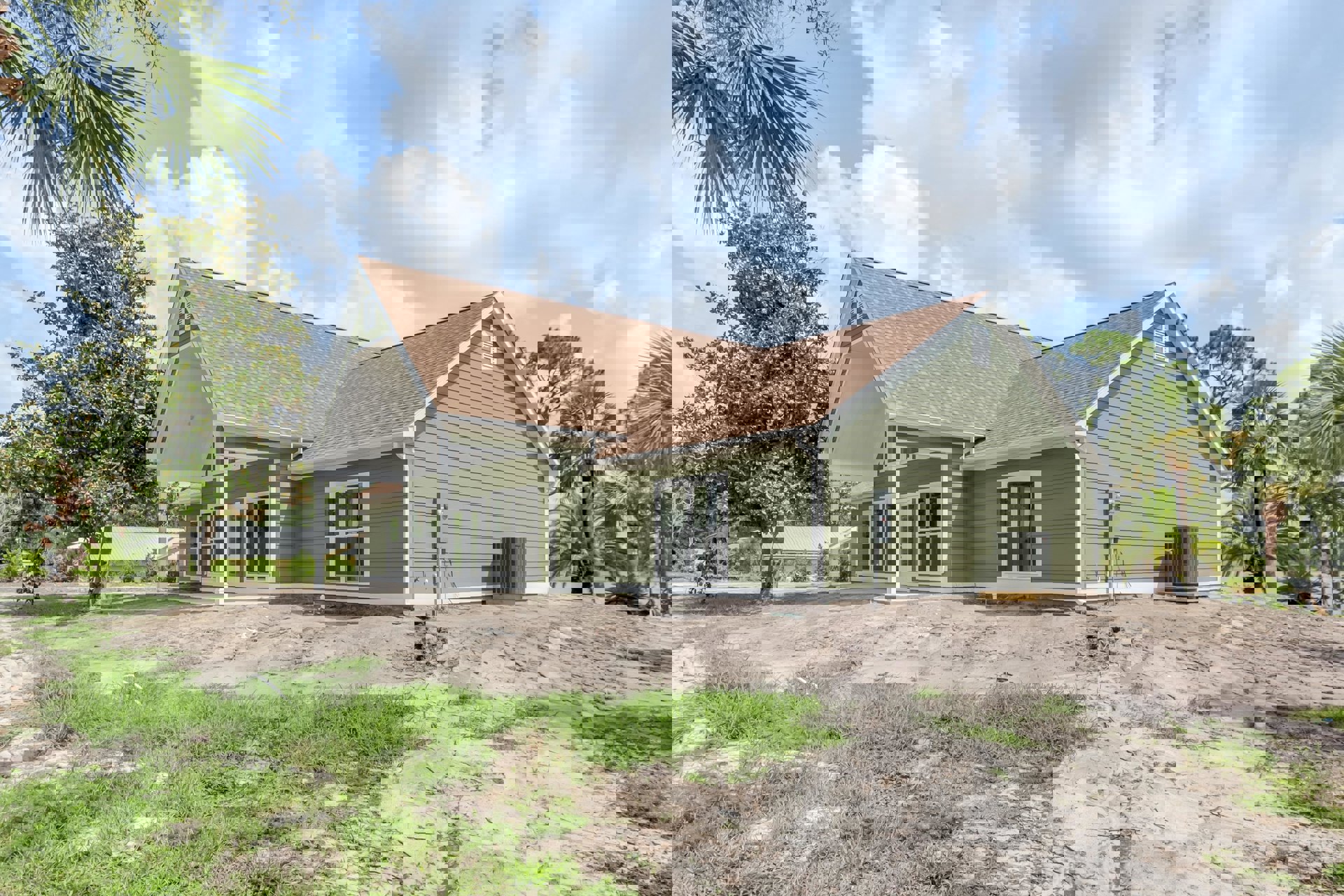 White-sided house with large windows, surrounded by dirt patches and mature trees including leafy and palm varieties, under a partly cloudy sky.