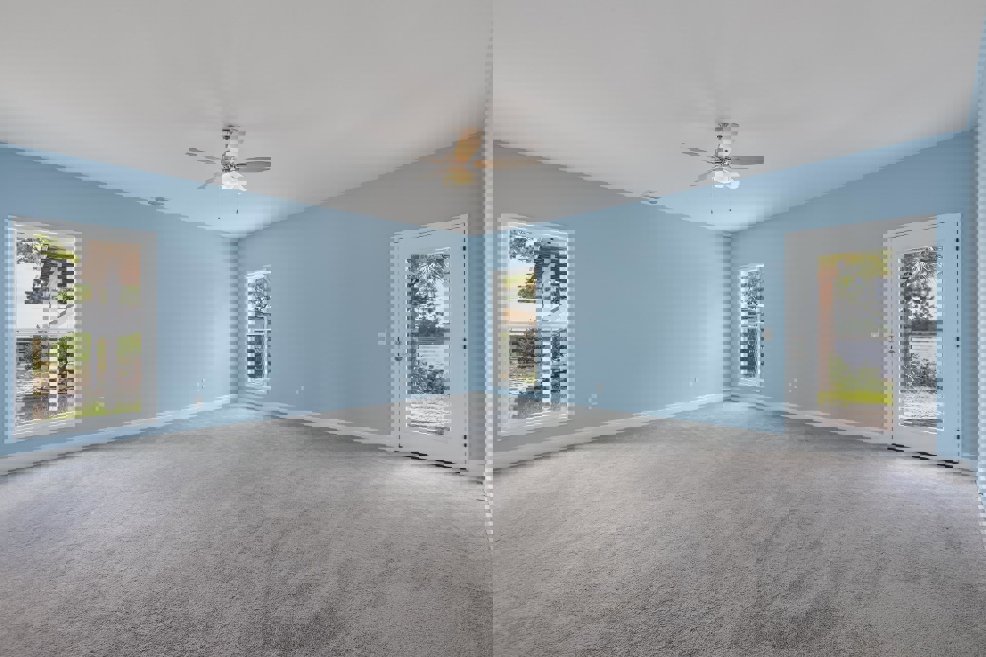 Carpeted bedroom with blue walls, ceiling fan with light, large window overlooking neighboring house, white door with glass panel, tree and water visible outside