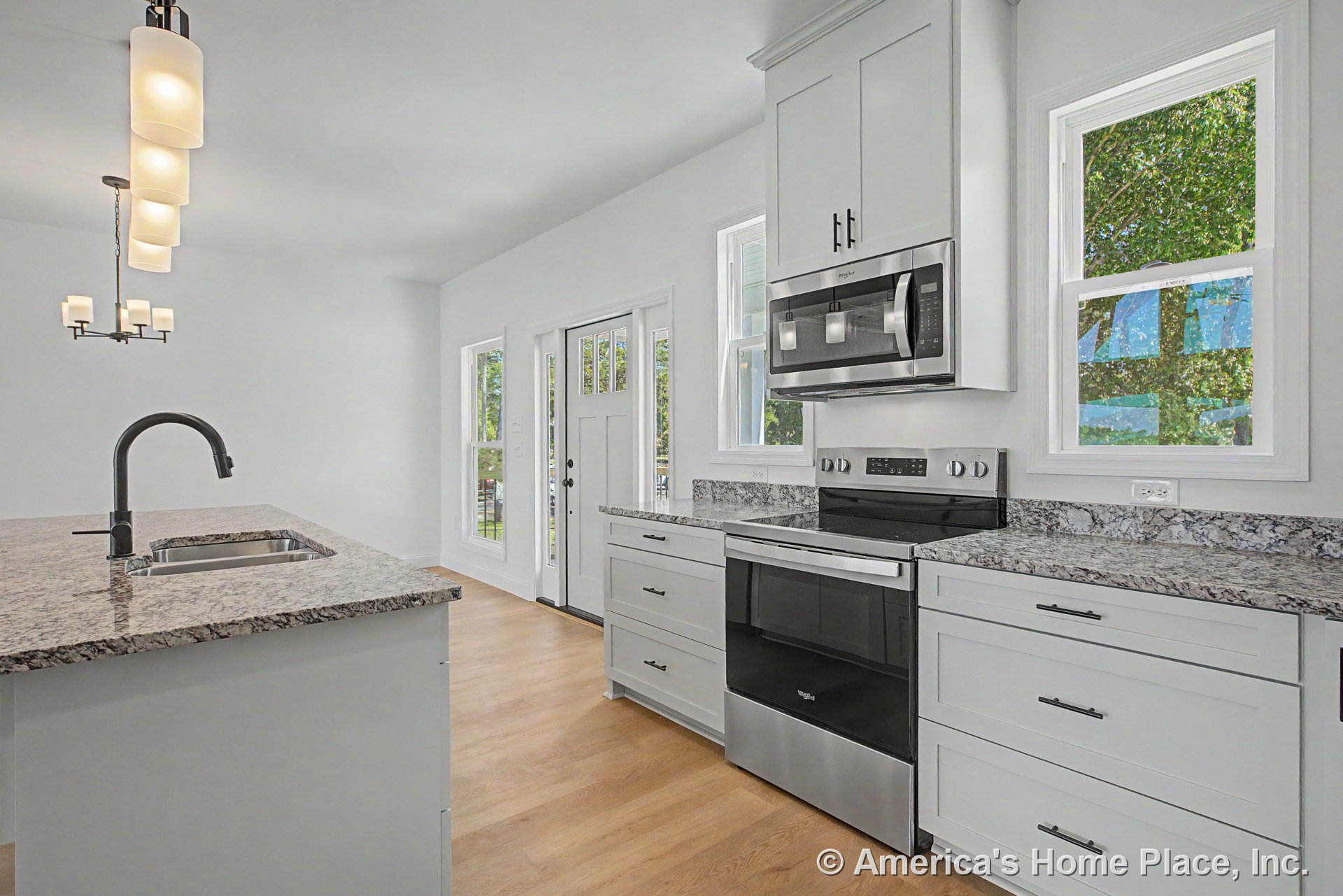 Granite island with undermount sink and matte black faucet, stainless steel range and microwave, white shaker cabinets, wood flooring, multiple windows with white trim, modern