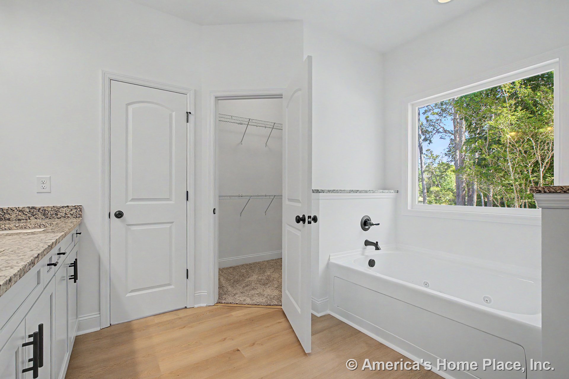 Built-in soaking tub with black fixtures positioned under a large window, granite vanity countertop, white shaker cabinets, walk-in closet with wire shelving, light wood flooring