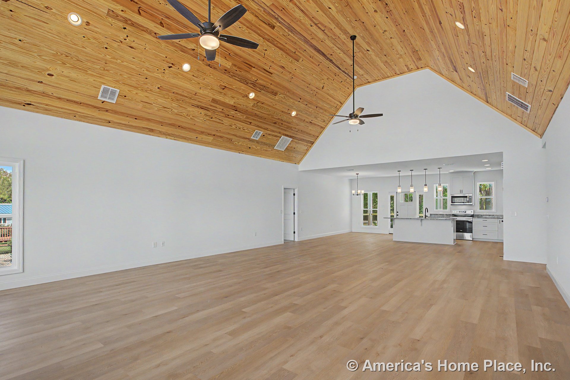 Vaulted wood plank ceiling spans open living area with light hardwood floors, modern kitchen featuring white cabinetry, pendant lights above island, and multiple large windows