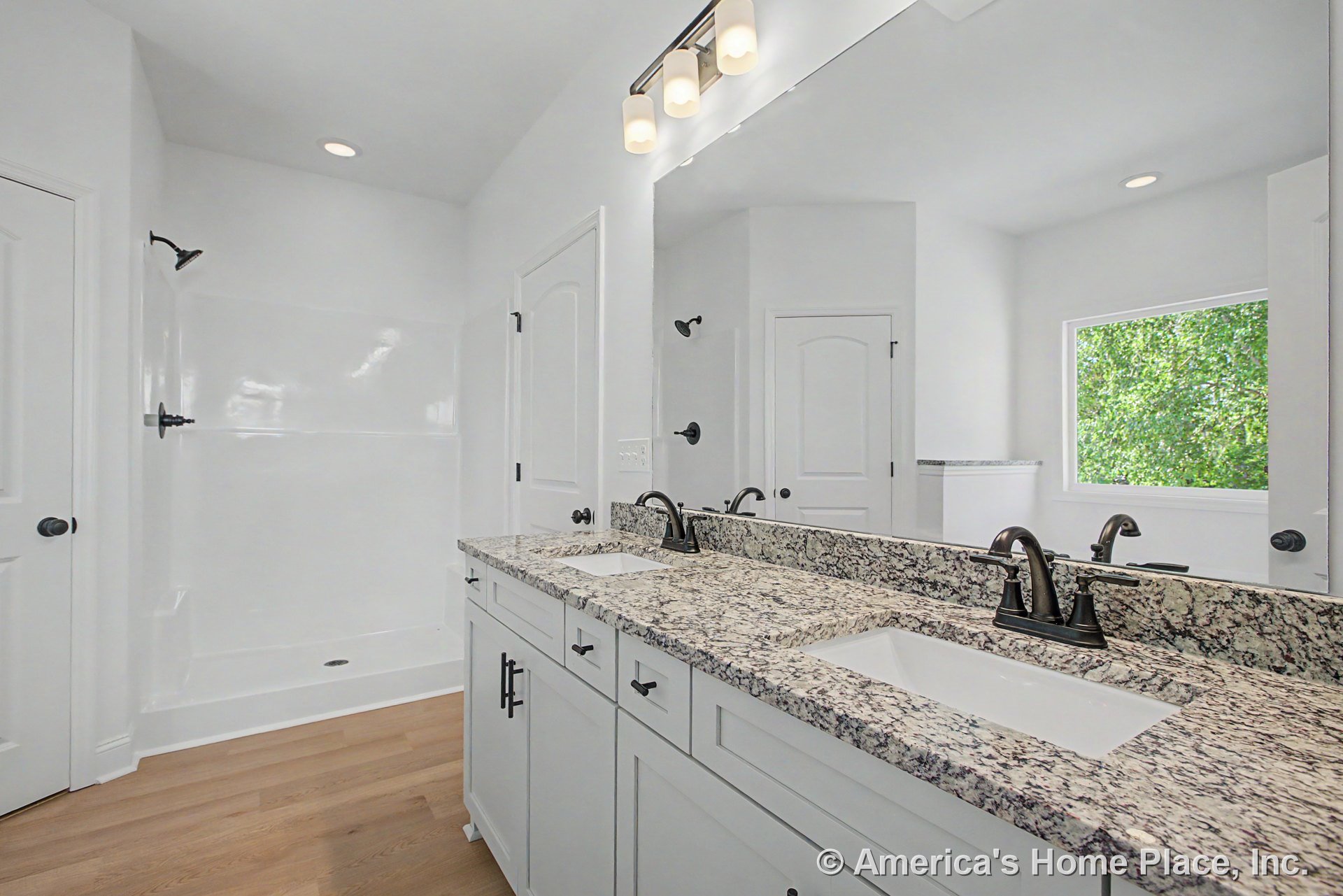 Double vanity with granite countertop, two undermount sinks, white shaker cabinets, walk-in shower with black fixtures, large wall mirror, recessed and vanity lighting, wood