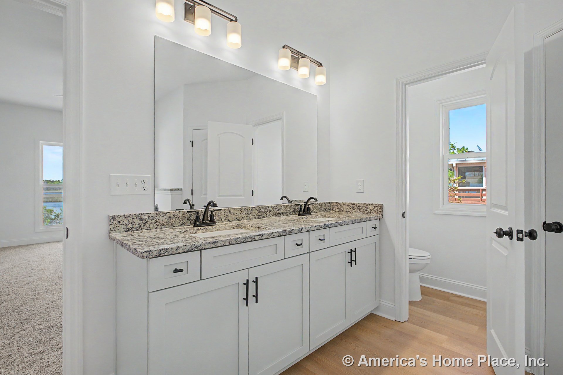 Double vanity with granite countertop, white shaker cabinets, large wall mirror, three-light vanity fixtures, wood flooring, window with trim, white door, chrome faucets, and