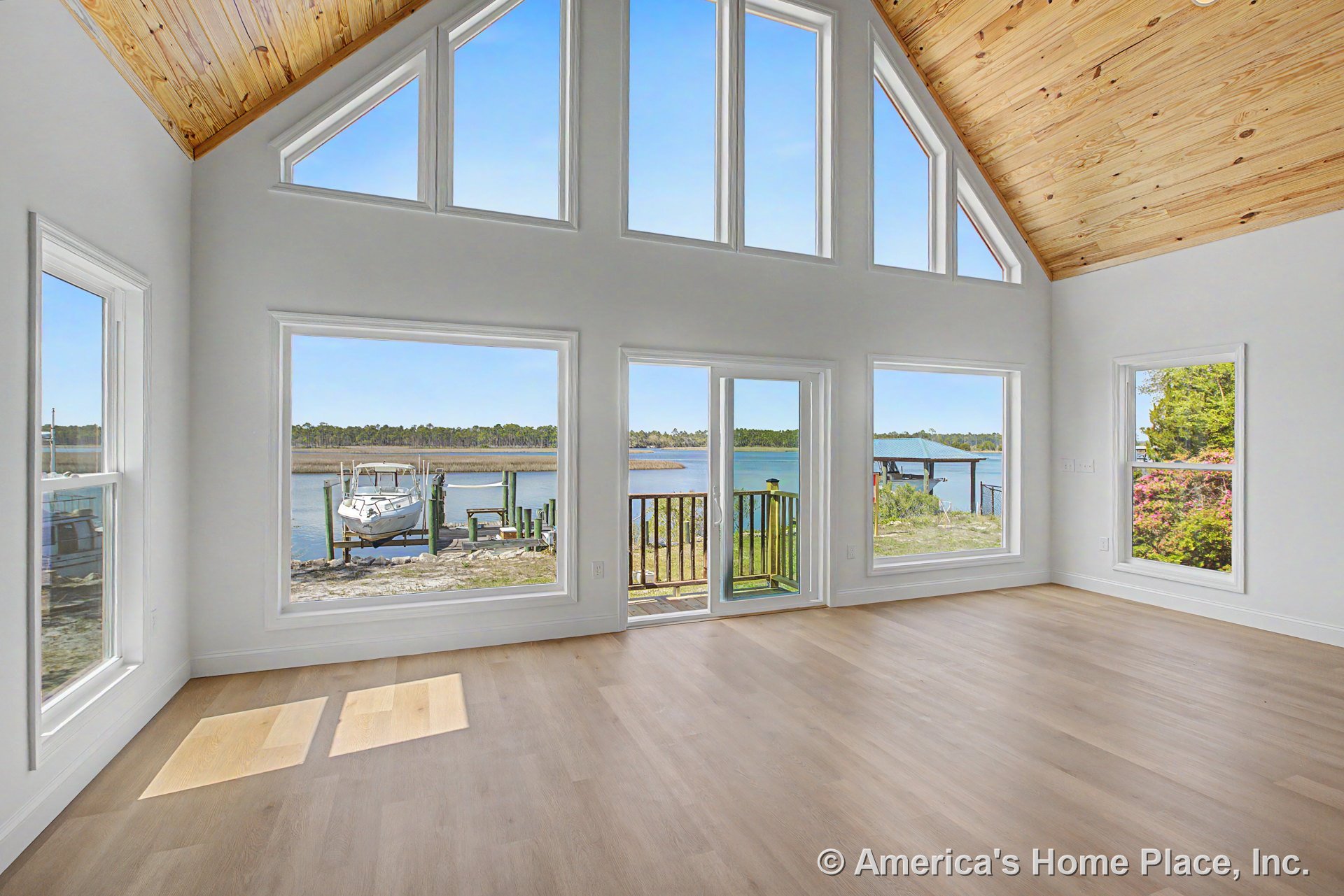 Vaulted wood plank ceiling and light wood flooring in an open living area with large picture windows, sliding glass door, white walls and trim, overlooking a waterfront view.