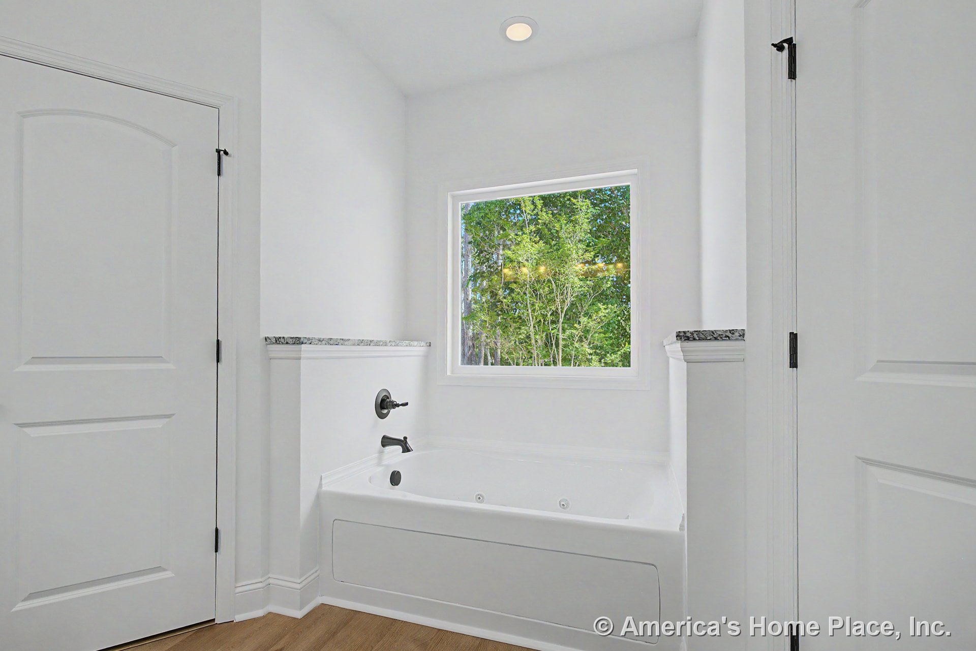 Built-in white soaking tub set below a large picture window with natural outdoor views, wood-look flooring, granite trim ledge, white paneled doors, recessed ceiling light, and