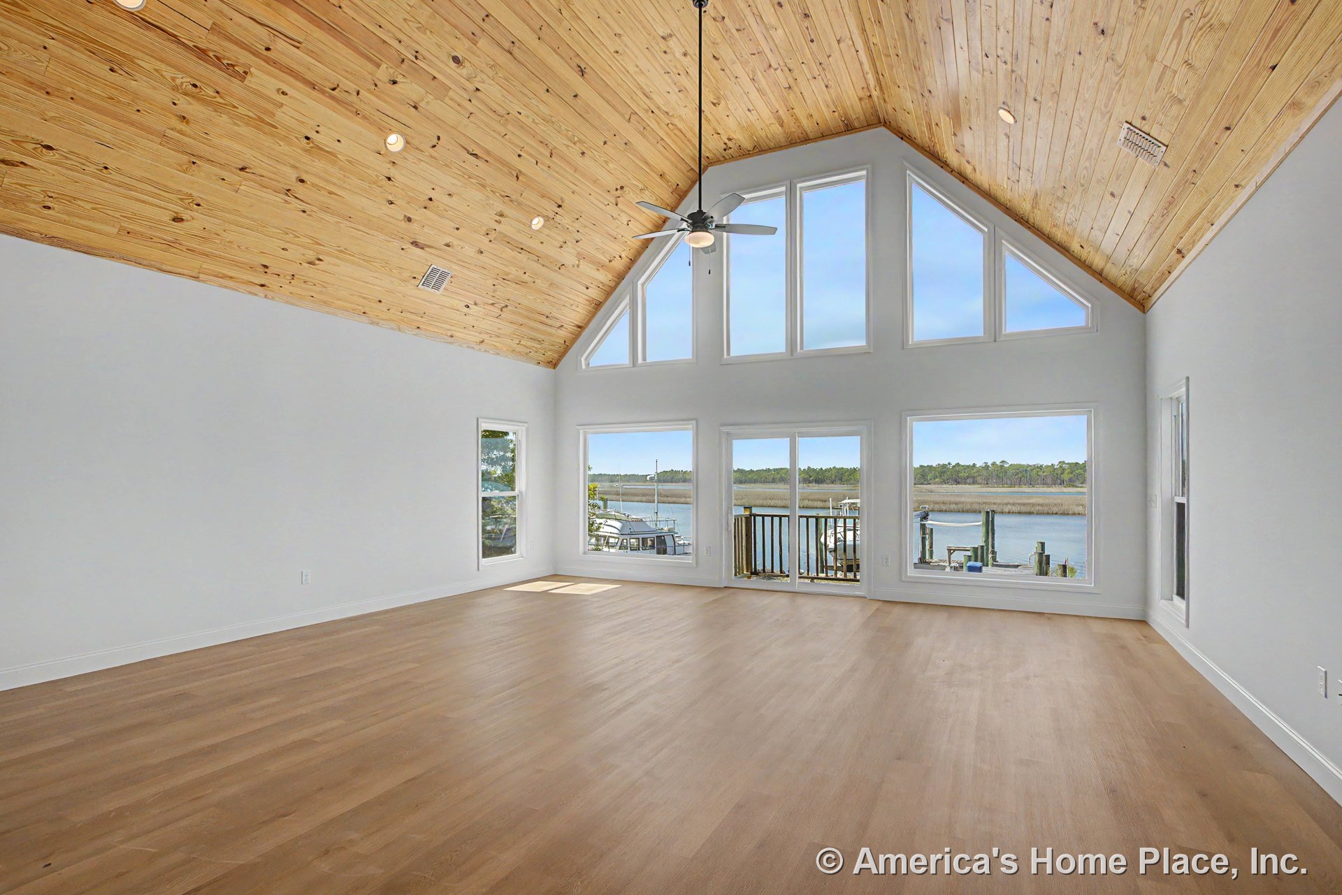 Vaulted wood plank ceiling with ceiling fan, large triangular windows and sliding glass door framed in white trim, light wood flooring, abundant natural light, waterfront view.