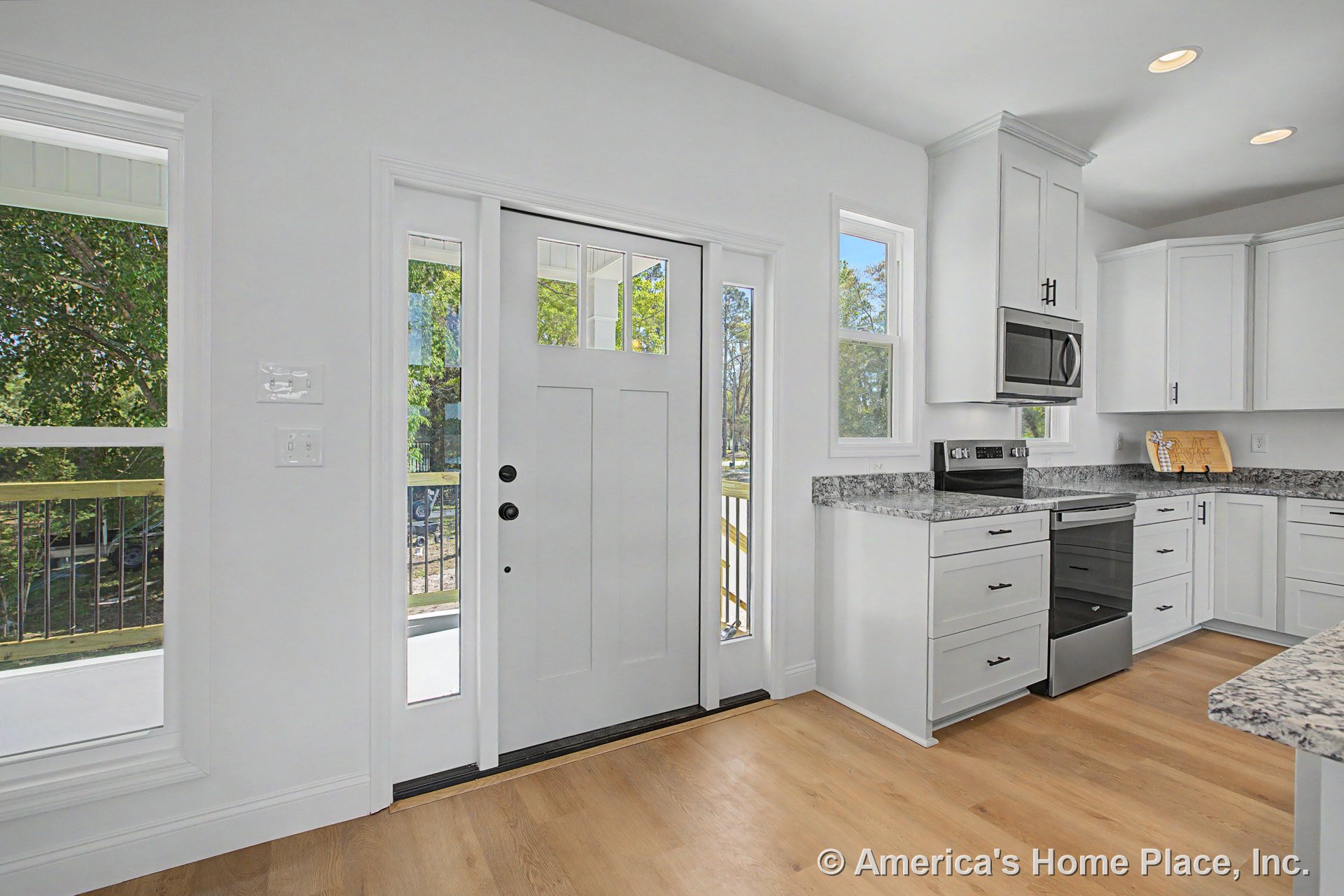 White shaker cabinets and granite countertops with stainless steel appliances in a kitchen next to a front entry door with sidelights, multiple large windows, light wood flooring