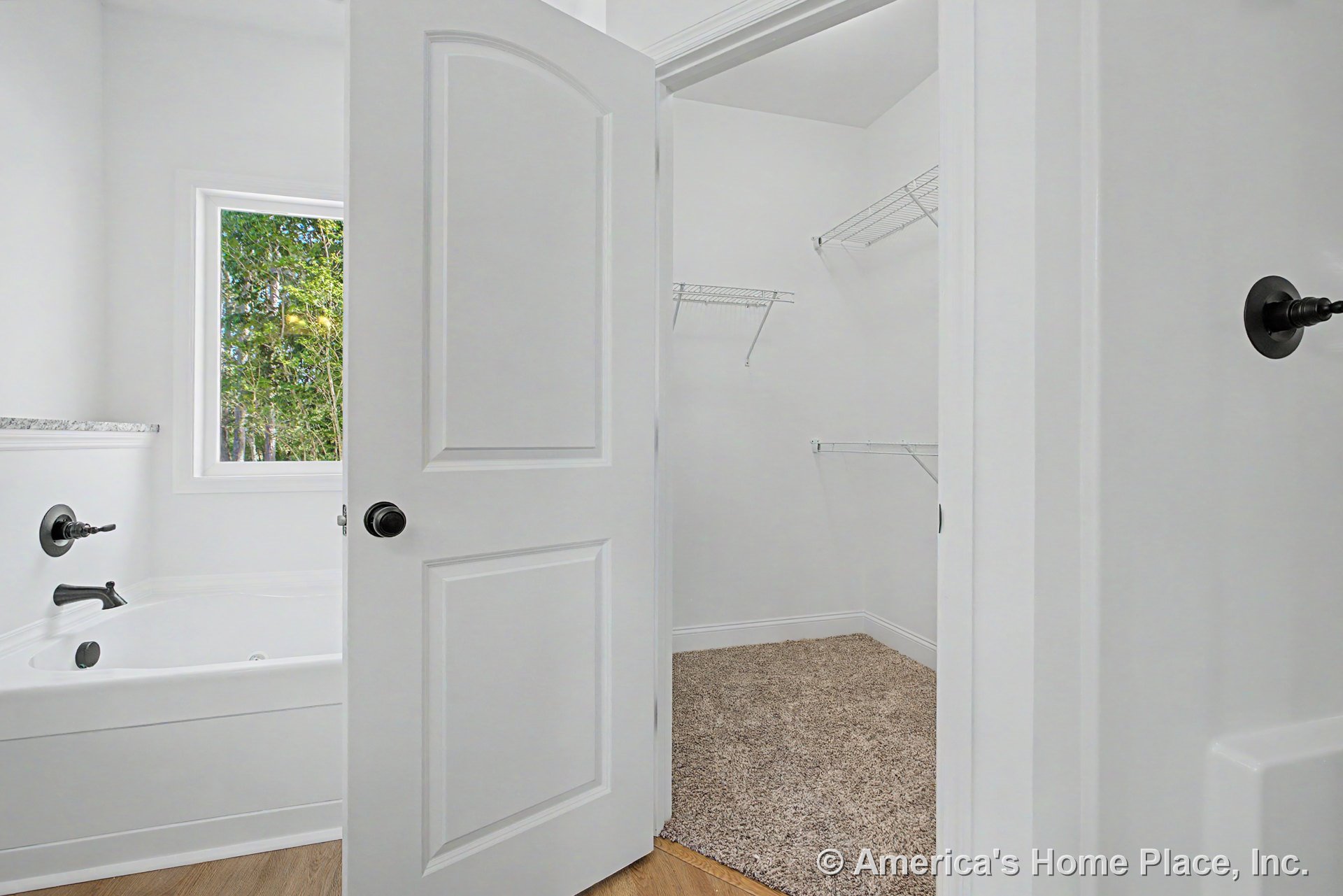 Soaking tub with black fixtures beneath a large window with white trim, adjacent walk-in closet featuring wire shelving and carpeted flooring, white paneled door, wood-look