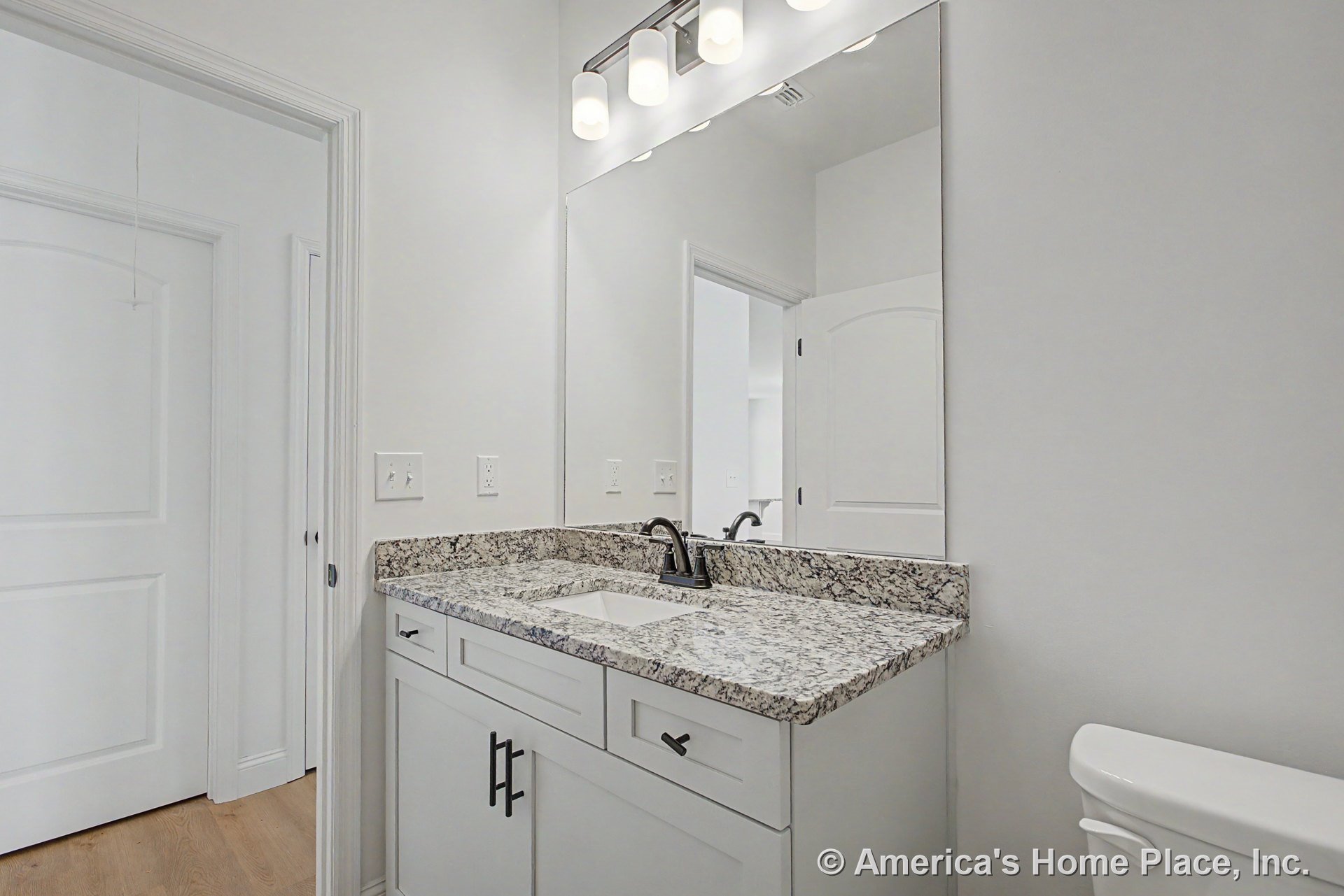 Granite vanity with undermount sink, white shaker cabinets, large wall mirror, three-light fixture above, and white paneled doors with modern hardware.