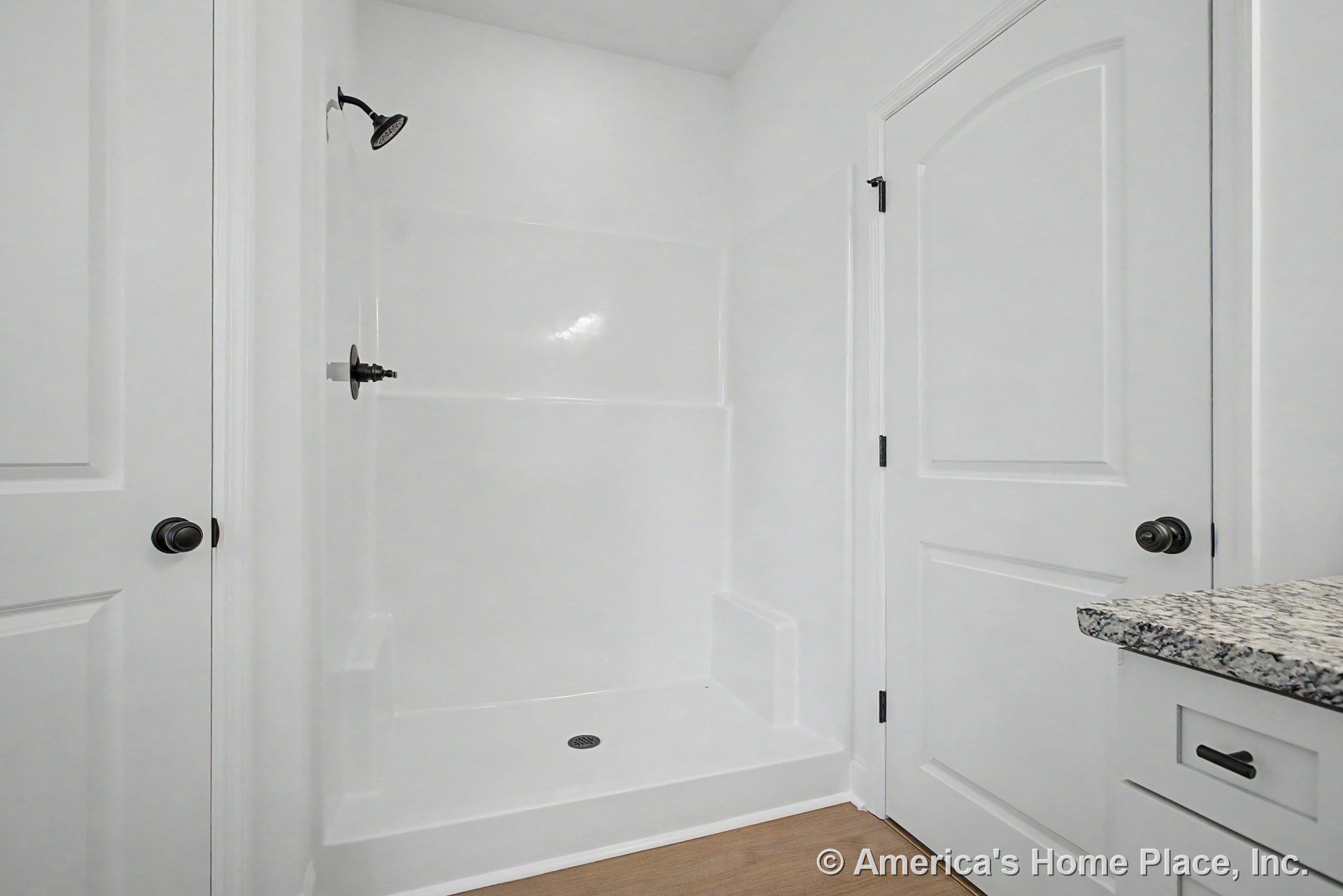 Walk-in shower with built-in bench, granite countertop, white paneled doors, wood-look flooring, black hardware, and white cabinetry.