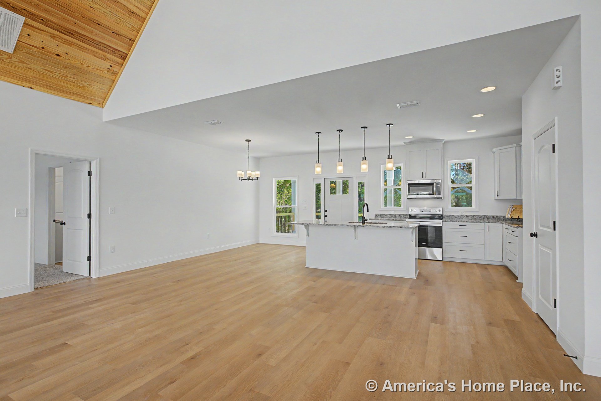 Open concept kitchen and living area with light wood plank flooring, white shaker cabinets, granite countertops, pendant lighting above island, vaulted ceiling with wood accent
